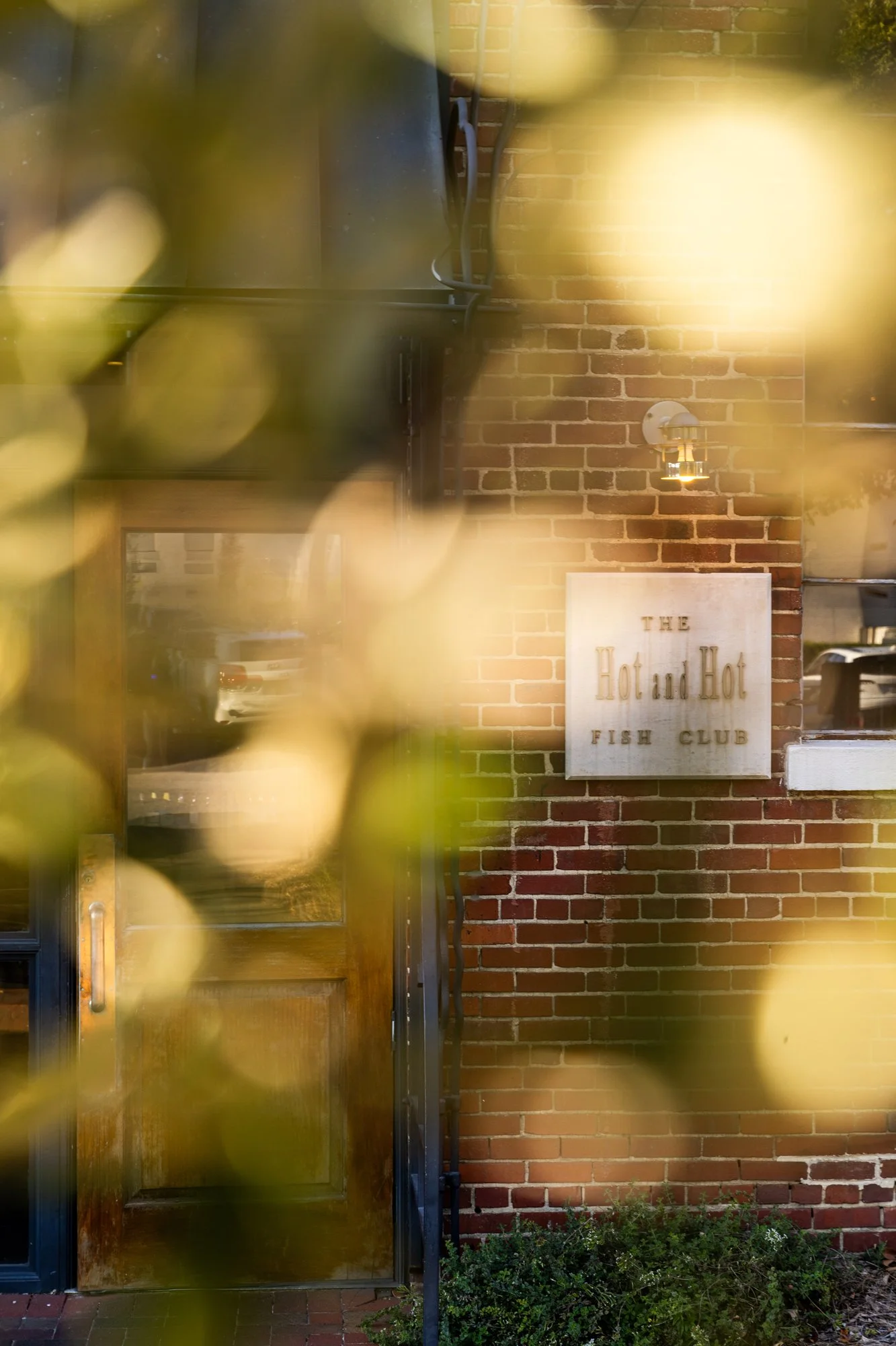 The front entrance of a restaurant called 'The Hot and Hot Fish Club' with a brick wall, a wooden door, and a sign near the door. Hot and Hot Fish Club Restaurant in Birmingham, Alabama.