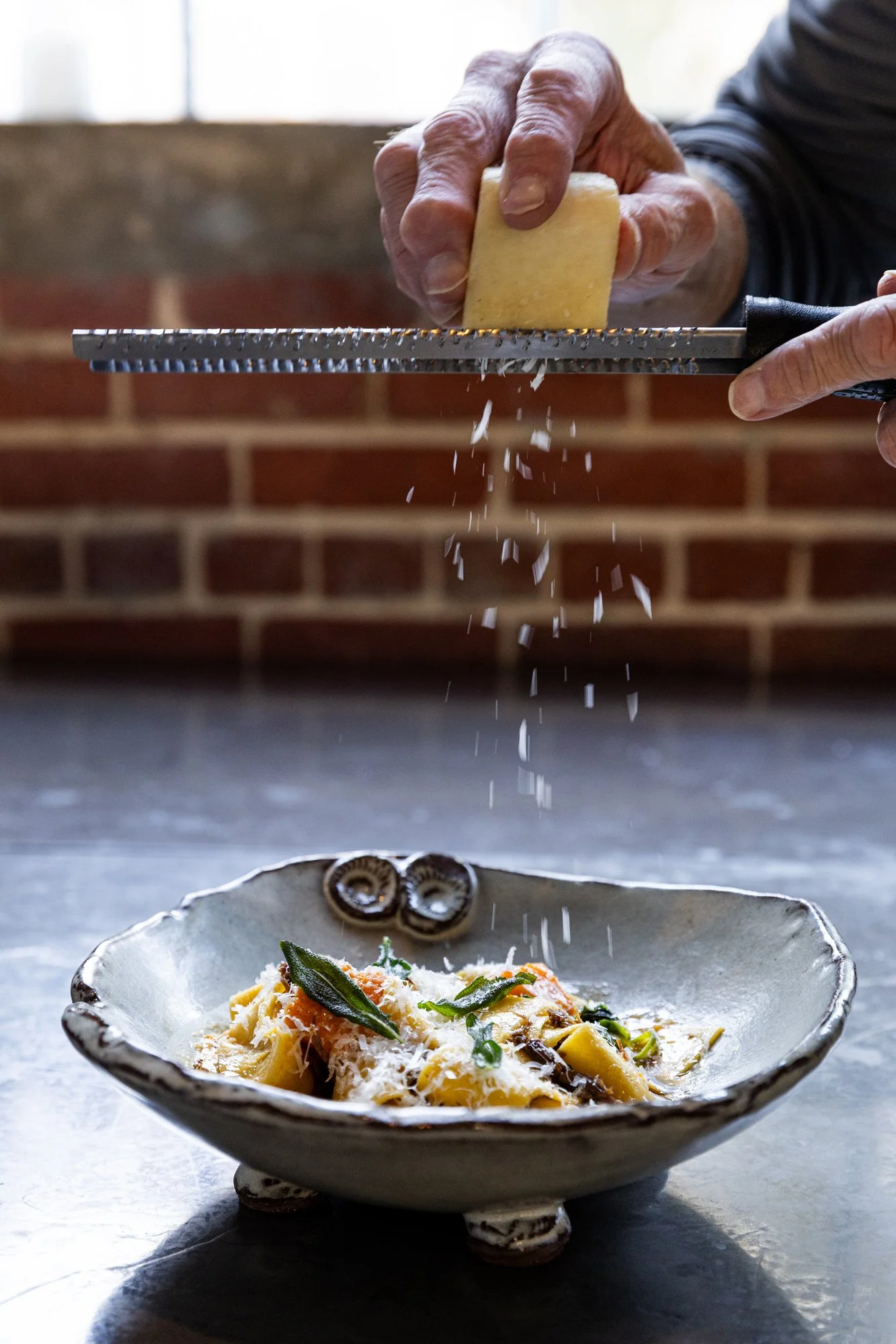 A person grating cheese over a plate of pasta with basil and cheese, with a brick wall in the background. Hot and Hot Fish Club Restaurant in Birmingham, Alabama.