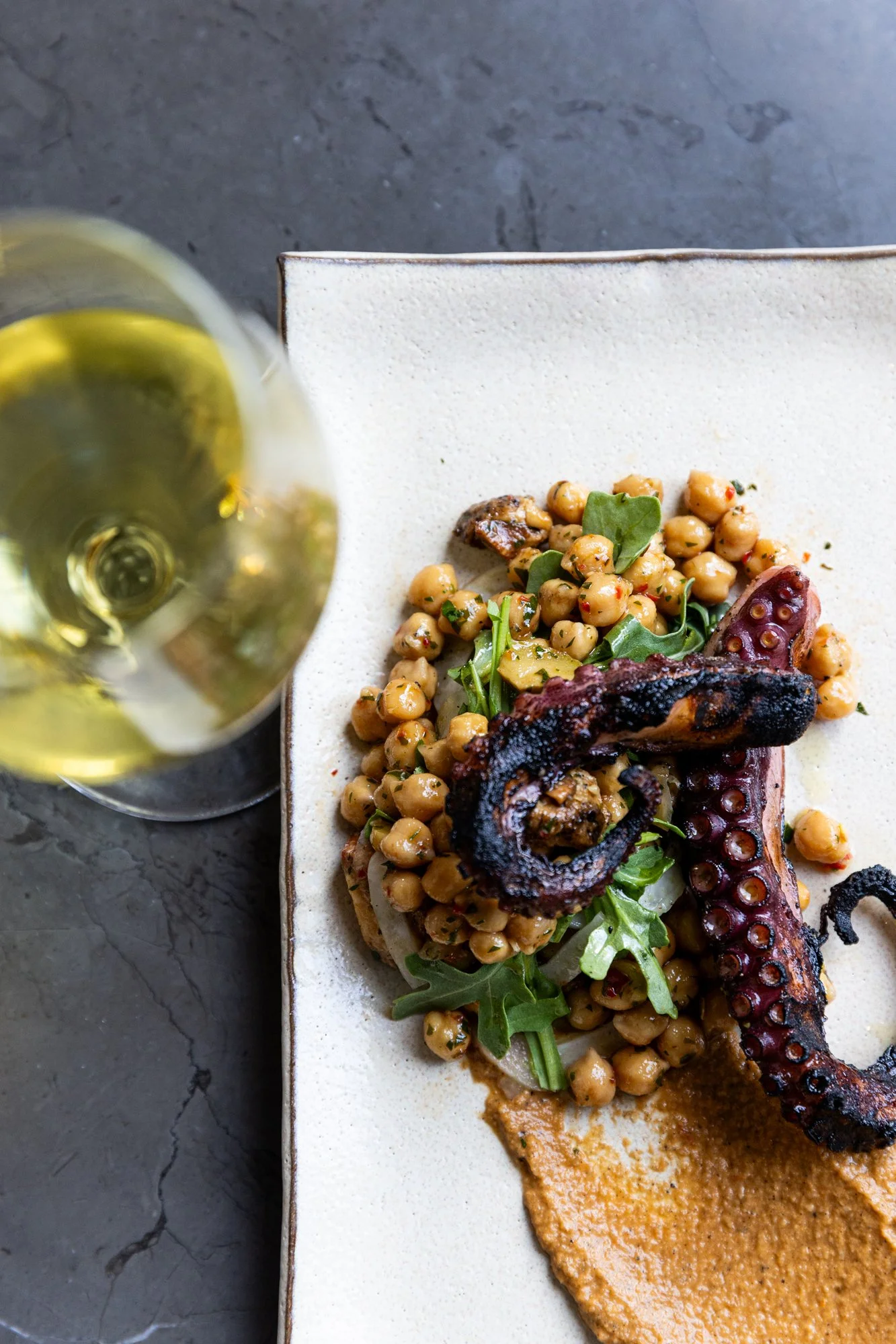Grilled octopus tentacle with chickpeas, greens, and sauce on a white plate, with a glass of white wine. Hot and Hot Fish Club Restaurant in Birmingham, Alabama.