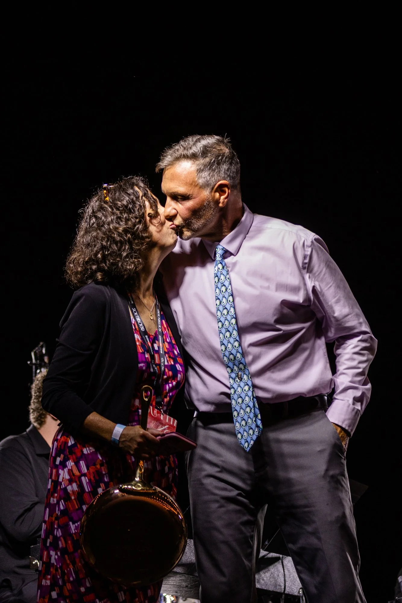A man and woman share a kiss on stage, with dark background and stage lighting. Southbound Food Festival in Birmingham, Alabama.