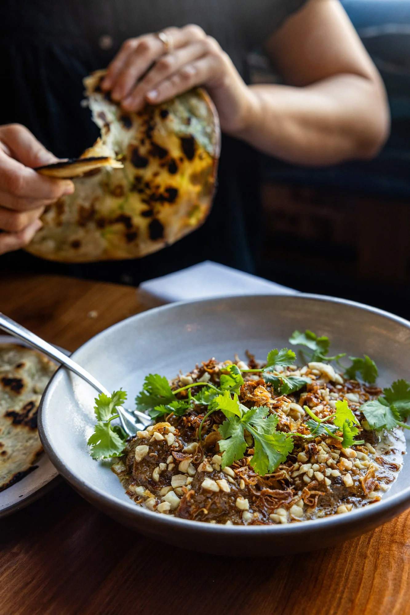 Person holds a piece of naan bread above a bowl of Mexican dish with cilantro, nuts, and shredded meat, on a wooden table.