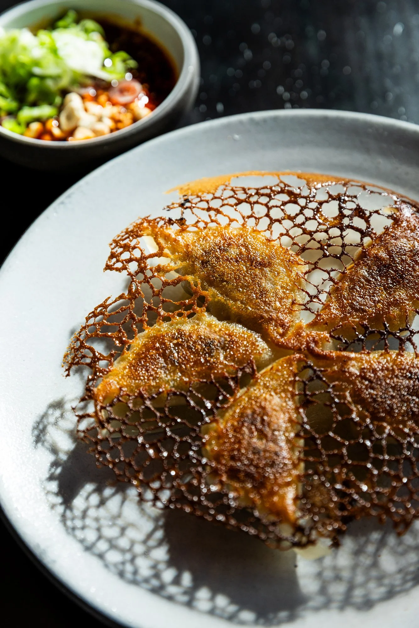 Close-up of four fried dumplings with a crispy, lacy, lattice-like surface on a gray plate, with a small bowl of dipping sauce with chopped green onions in the background.