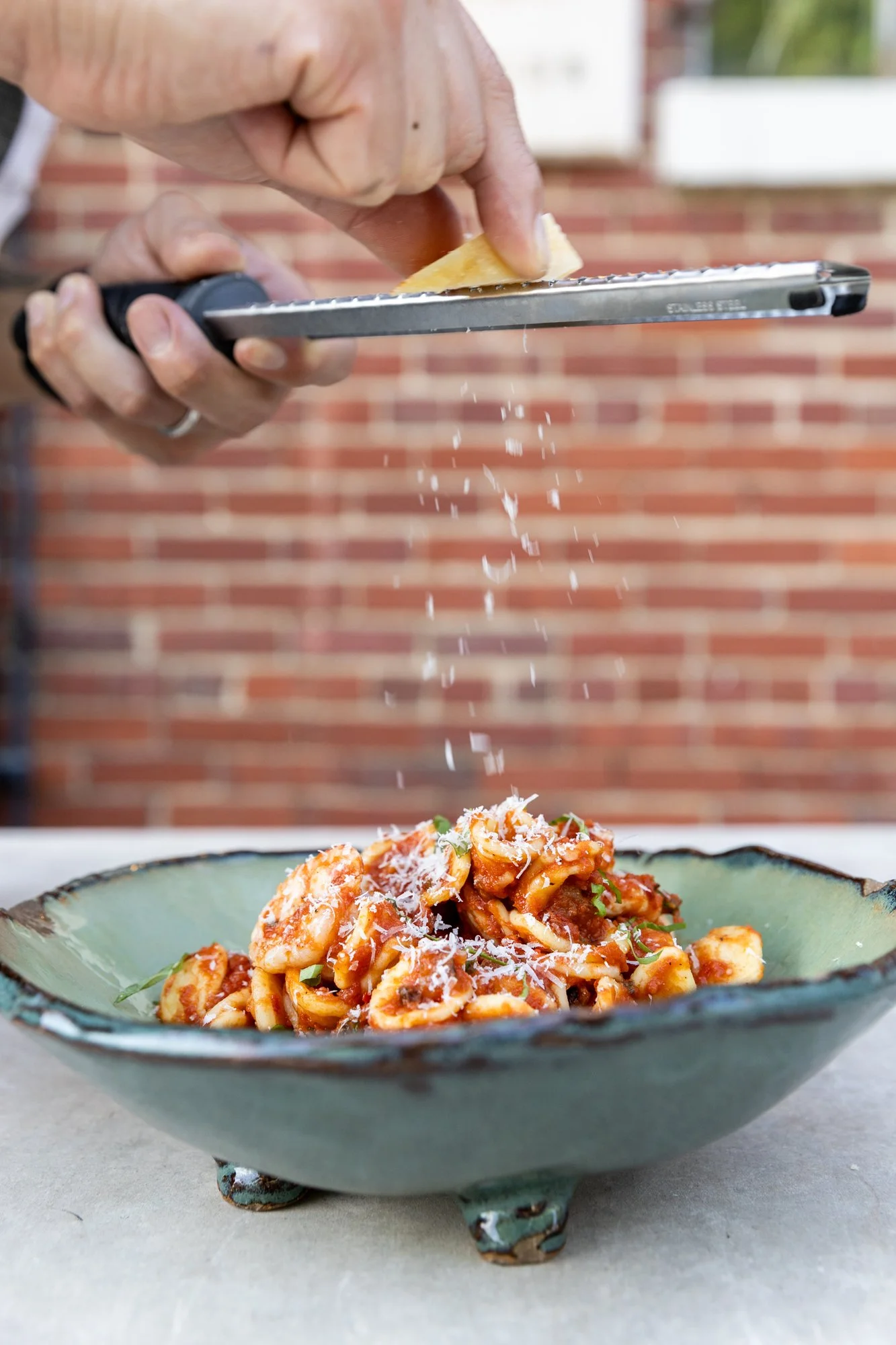 Person grating cheese over a bowl of pasta with tomato sauce on a table in front of a brick wall. Hot and Hot Fish Club Restaurant in Birmingham, Alabama.