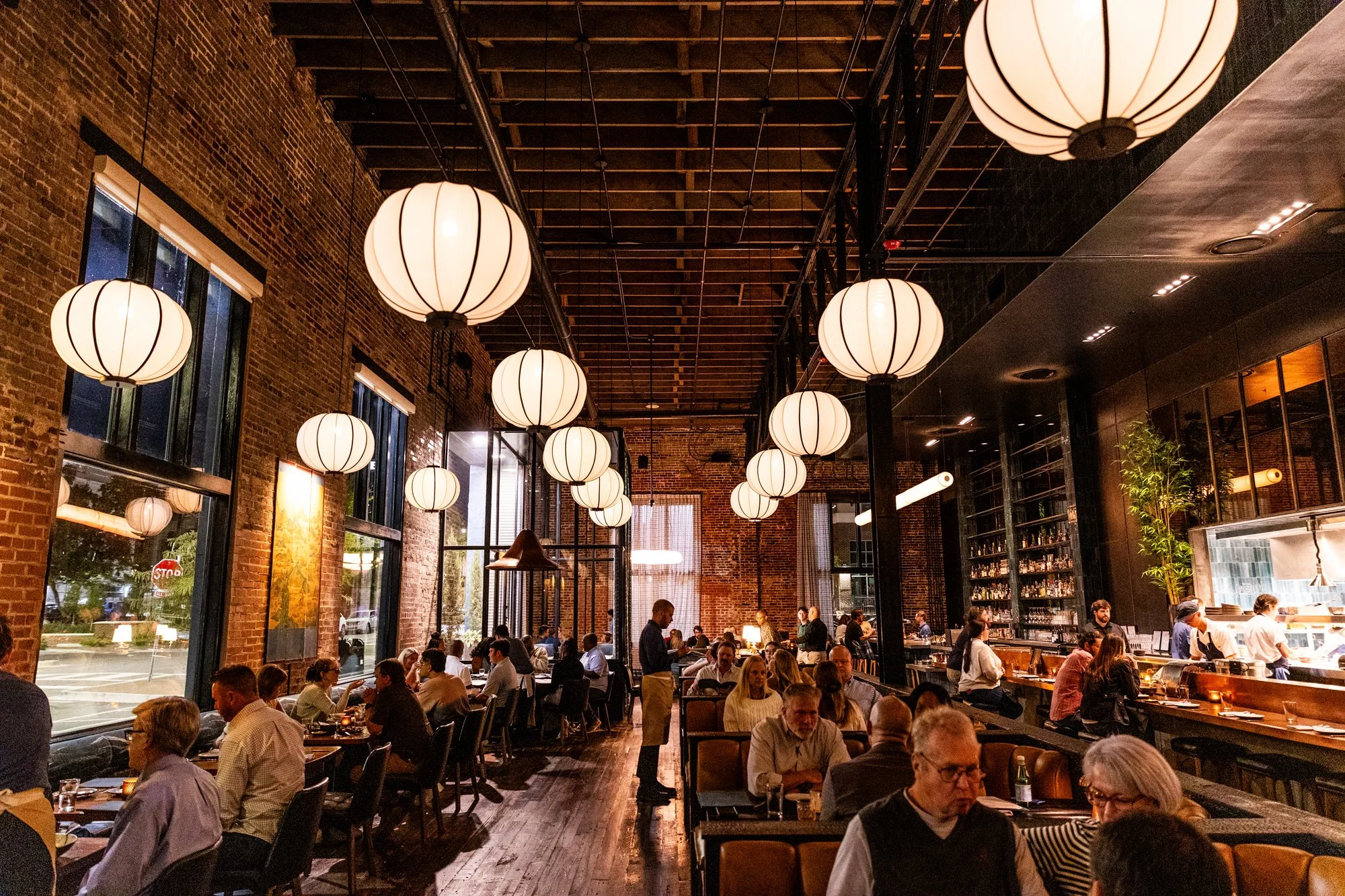 Interior of a busy restaurant with hanging paper lantern lights, brick walls, large windows, and a bar area with staff preparing food.