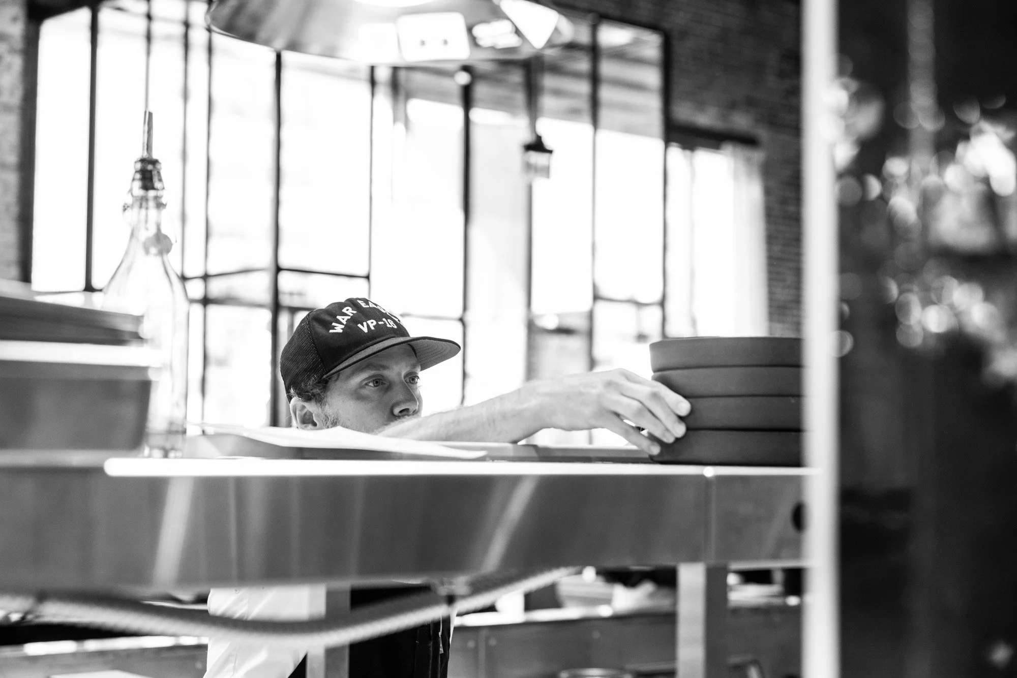 A person wearing a cap labeled 'WAR' and 'VP-23' is reaching to place or pick up pizza boxes on a shelf in a restaurant or kitchen.