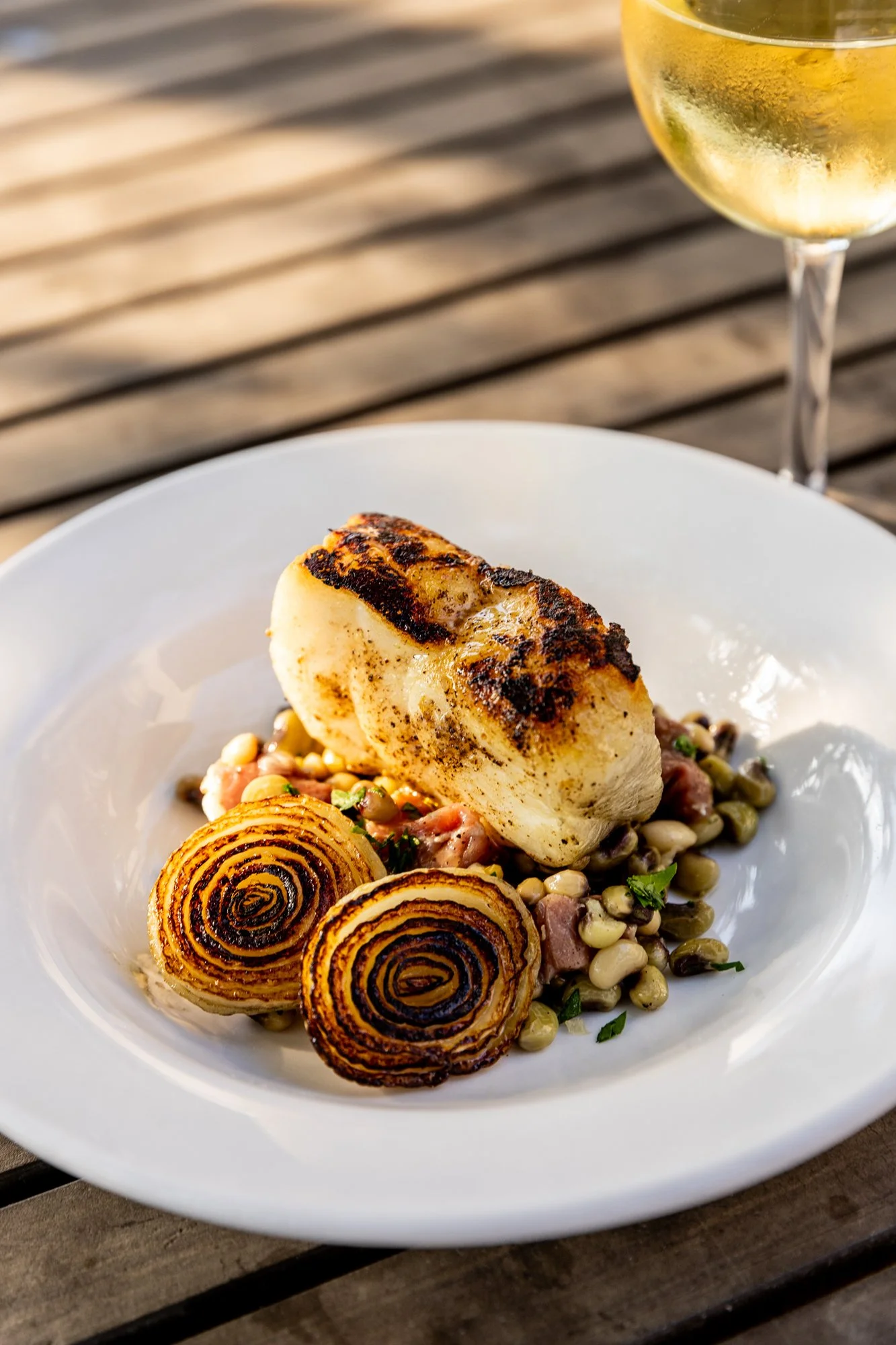 A plated dish with a grilled piece of fish, roasted onions, and a mixed bean salad, with a glass of white wine in the background. Ovenbird Restaurant in Birmingham, Alabama.