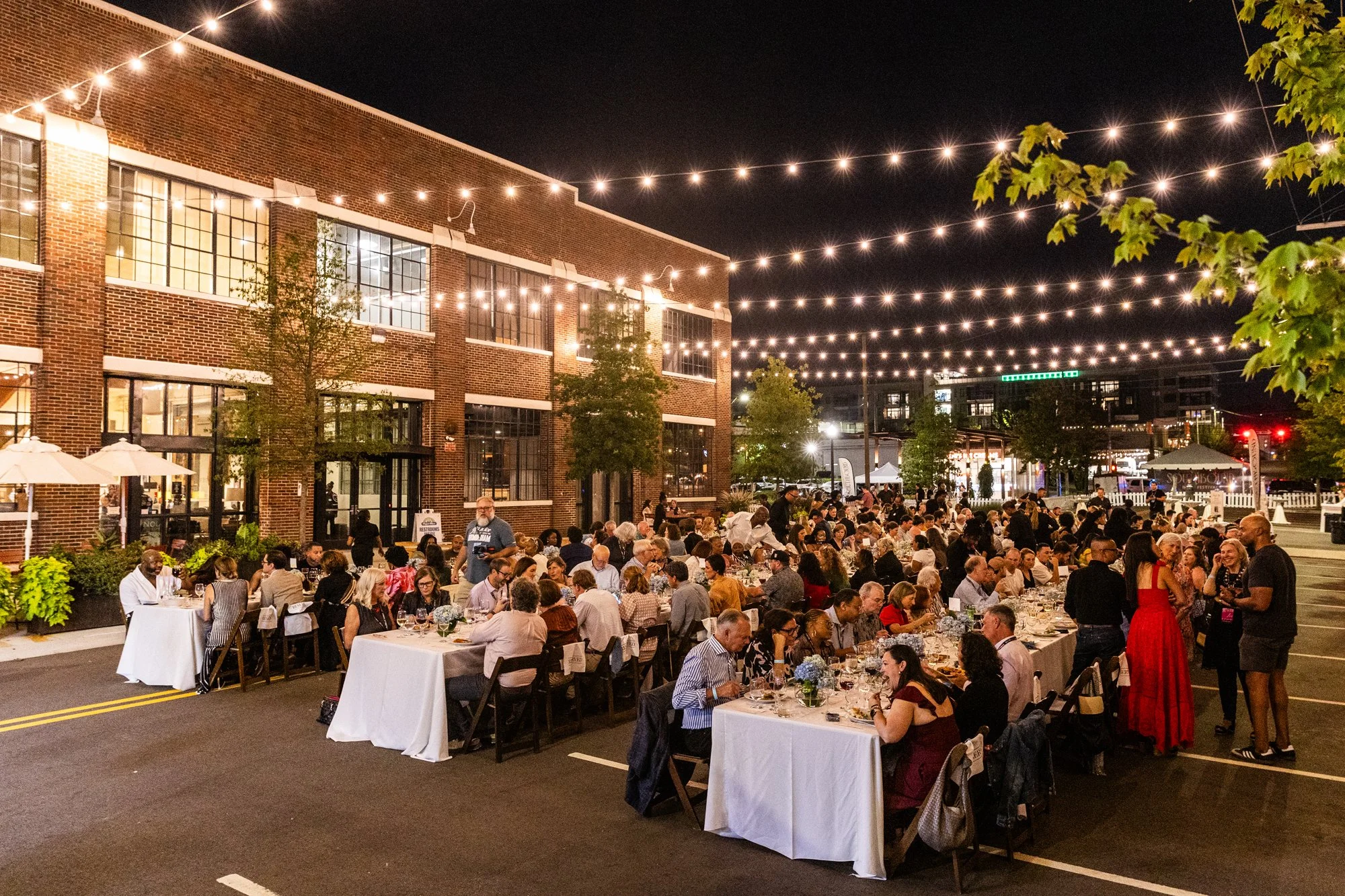 Outdoor evening dinner event with many people seated at long tables with white tablecloths, under string lights, outside a brick building, with some standing and socializing Southbound Food Festival in Birmingham, Alabama.
