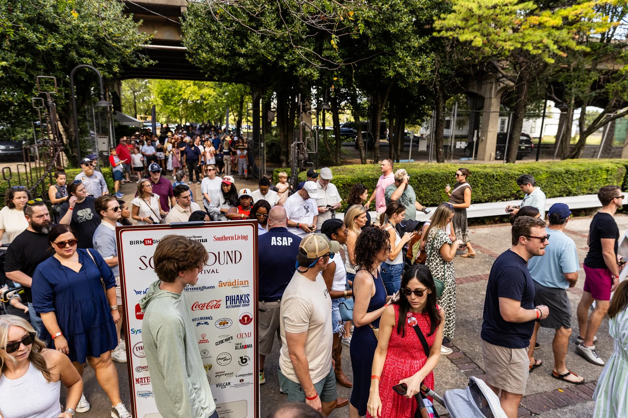Crowd of people gathering outdoors at a festival or event, some standing in line or walking, with a large sign visible in the foreground and trees overhead. Southbound Food Festival in Birmingham, Alabama.