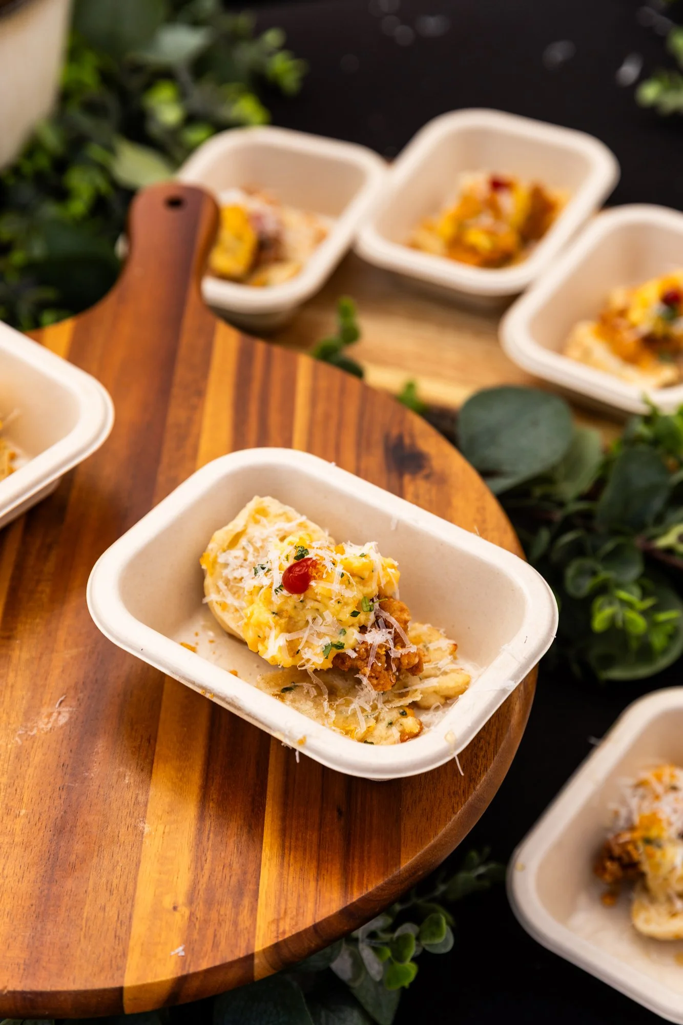 Creamy potato salad topped with shredded cheese, herbs, and a small red chili on a beige paper bowl, placed on a wooden board with more similar salads in the background. Southbound Food Festival in Birmingham, Alabama.