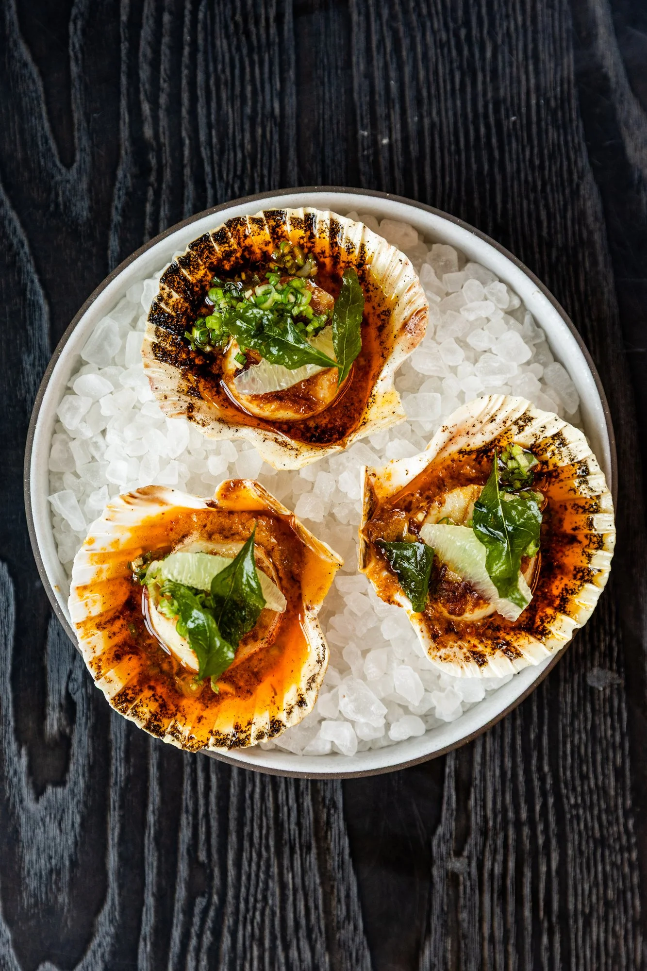 Three grilled scallops topped with herbs and sauce served on ice in a white bowl