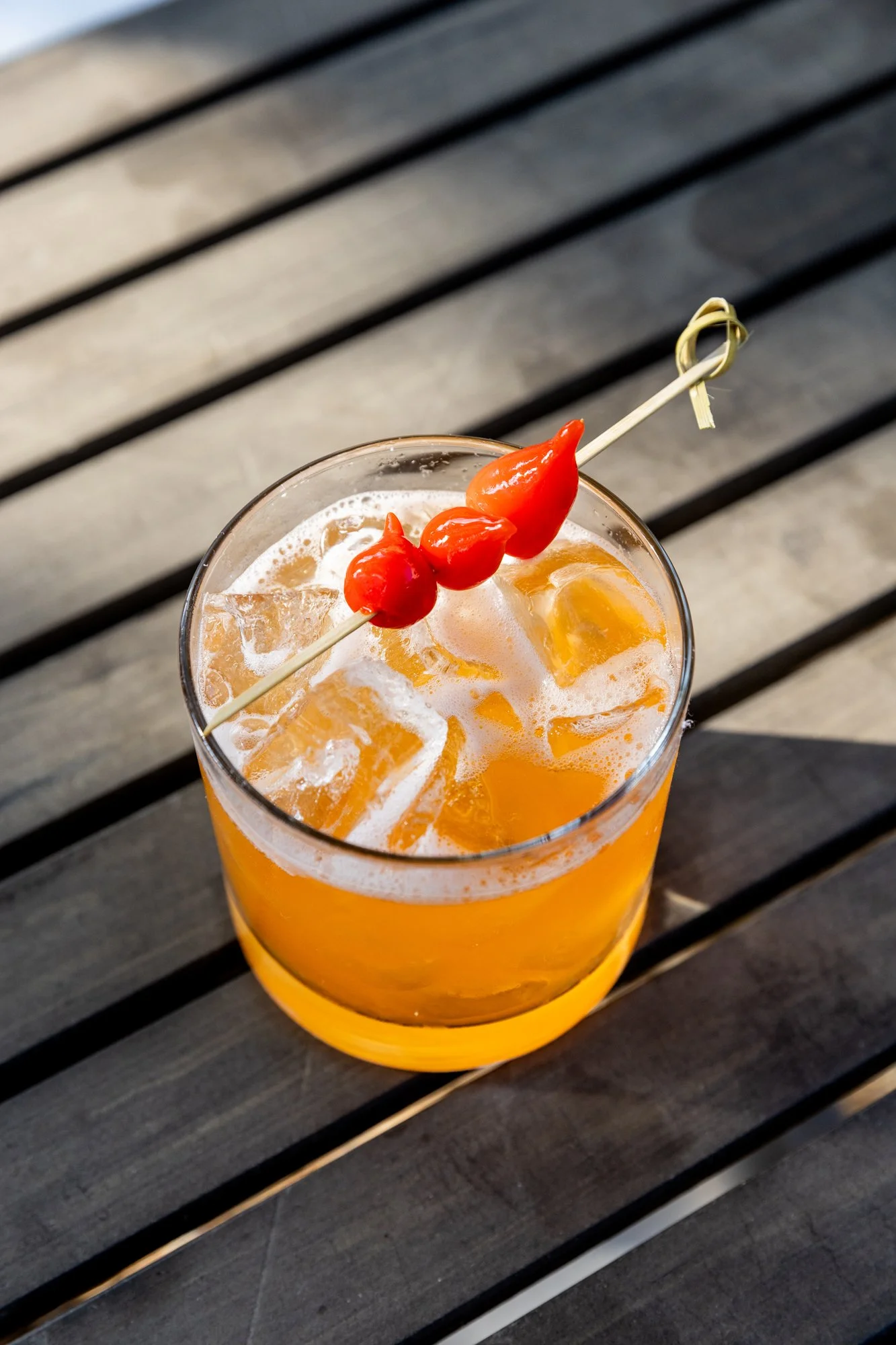 A glass of orange cocktail with ice and a garnish of red peppers on a wooden table. Ovenbird Restaurant in Birmingham, Alabama.