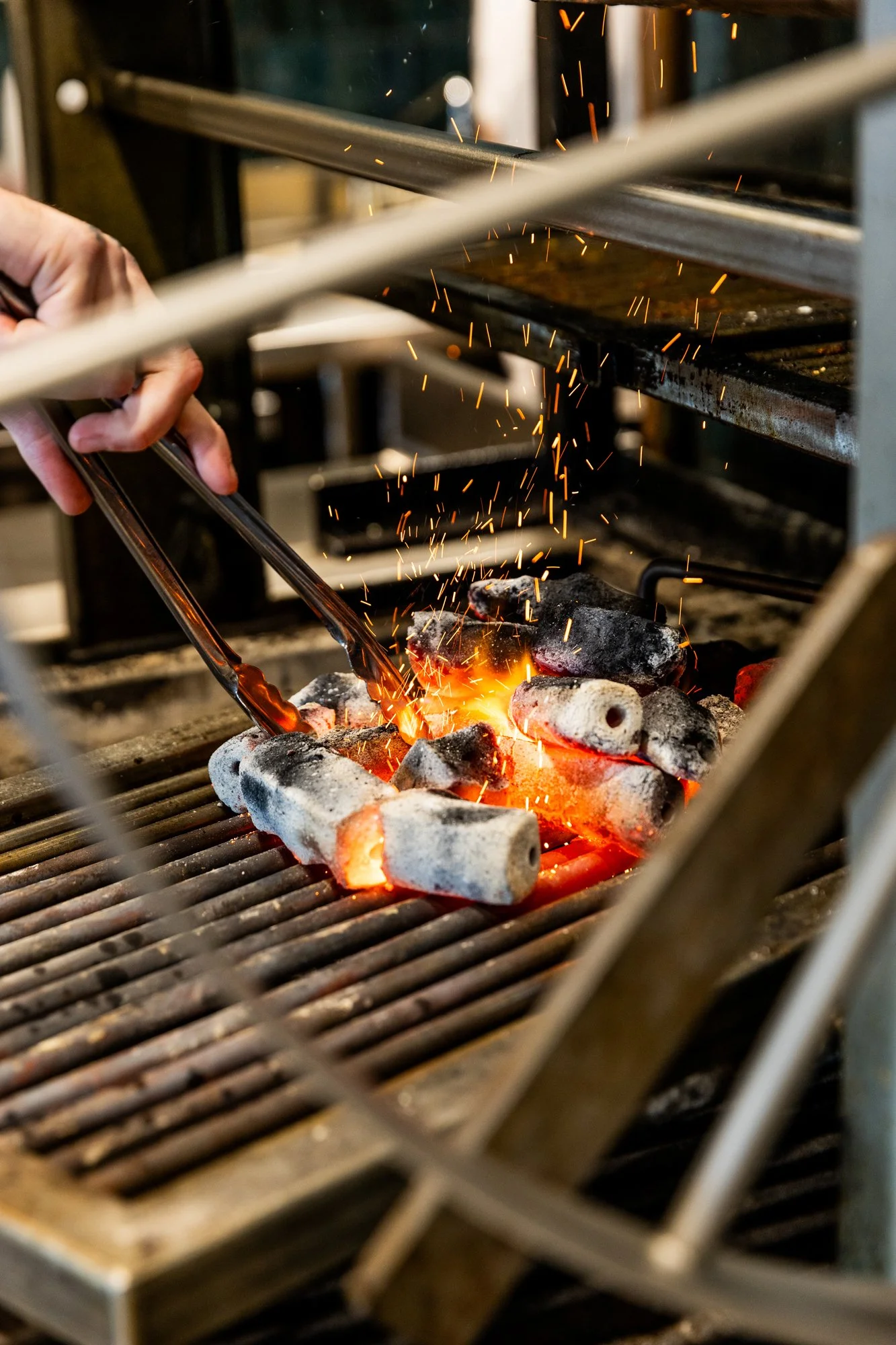A person using tongs to place red-hot metal pieces on a grill with glowing charcoal and sparks flying.