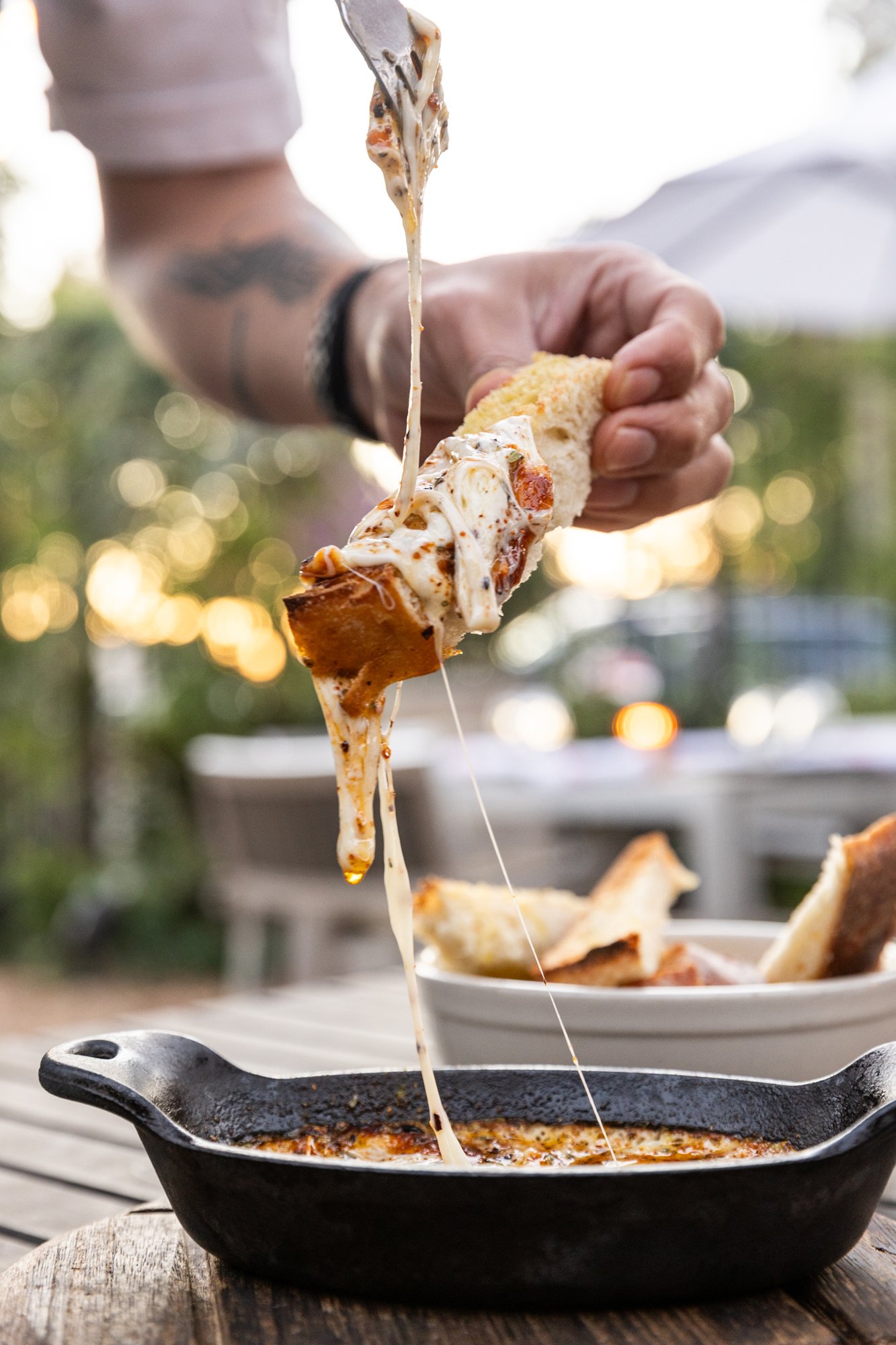 Person holding a slice of bread with melted cheese and toppings, lifting it from a skillet of melted cheese and sauce. Ovenbird Restaurant in Birmingham, Alabama.