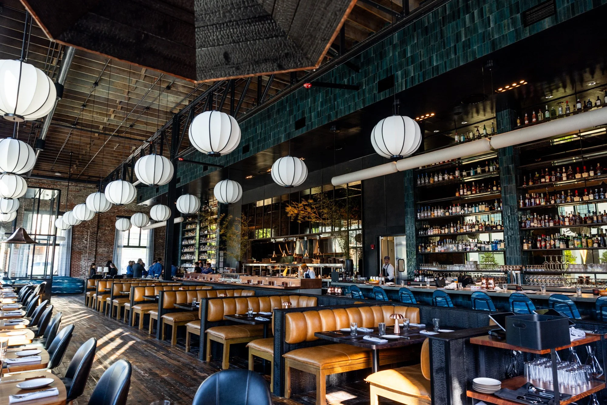 Interior of a modern restaurant with hanging white lantern lights, wooden floors, leather booths, and a bar with shelves of bottles.