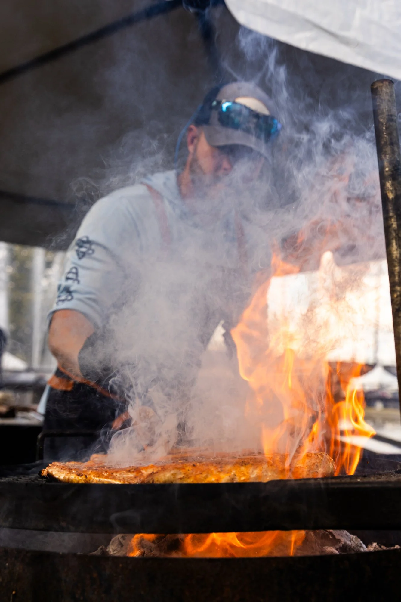 Man grilling meat over an open flame with smoke and fire visible, wearing sunglasses and a cap. Southbound Food Festival in Birmingham, Alabama.
