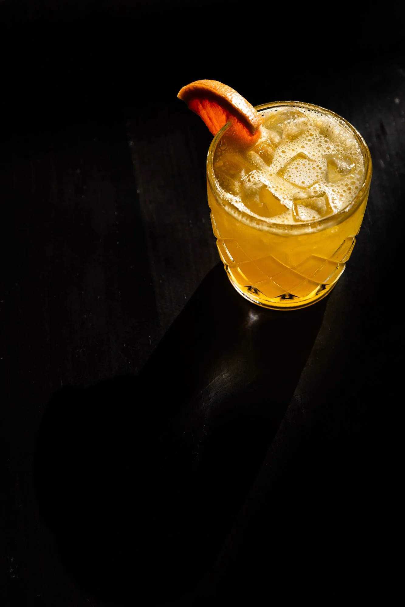 A yellow cocktail with ice cubes and a slice of orange on the rim, served in a textured glass on a dark surface, casting a shadow.