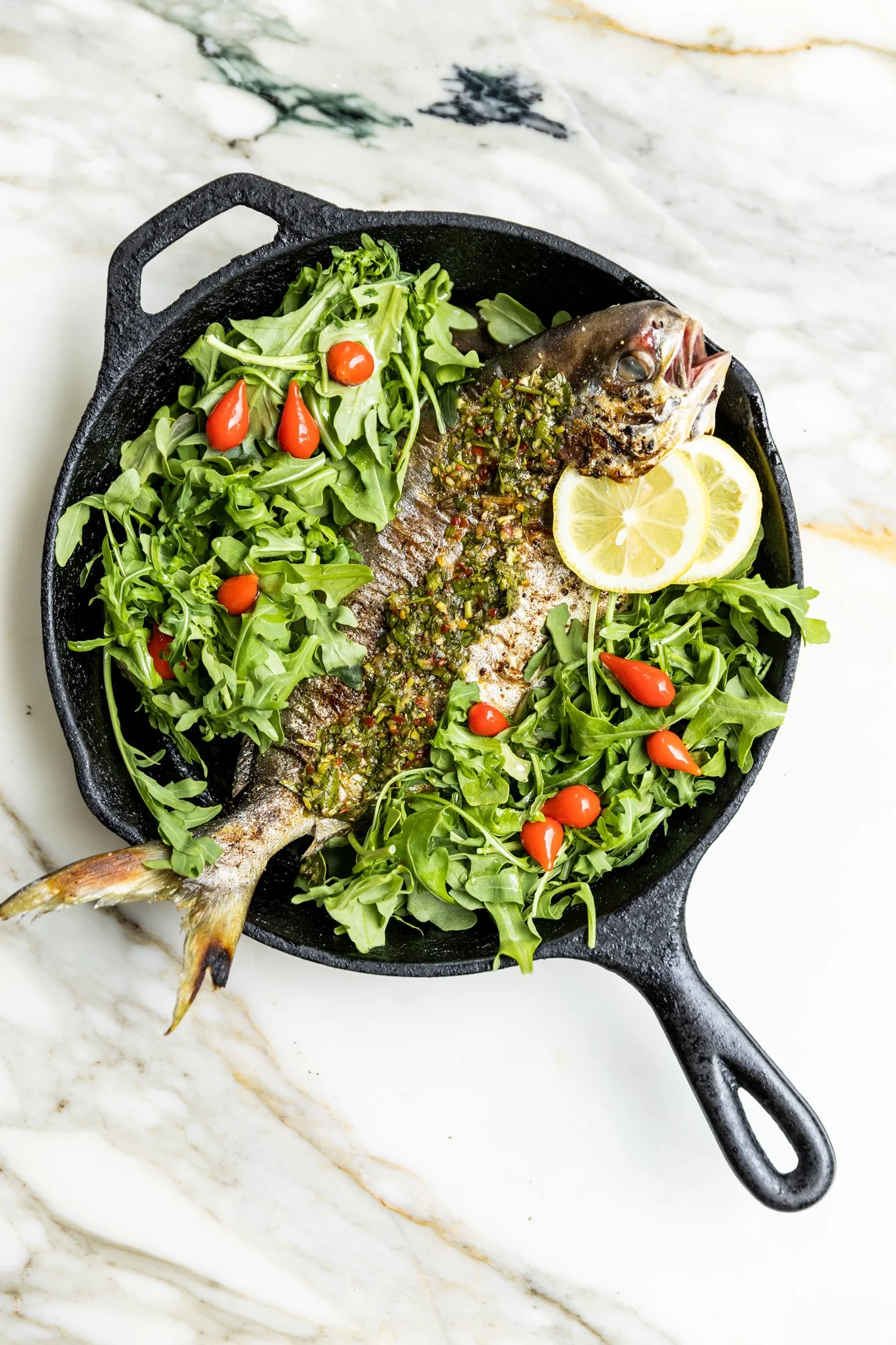 A whole fish garnished with herbs and chili flakes, served with lemon slices, and surrounded by green leafy vegetables and small red peppers in a black cast iron skillet on a white marble surface. Ovenbird Restaurant in Birmingham, Alabama.