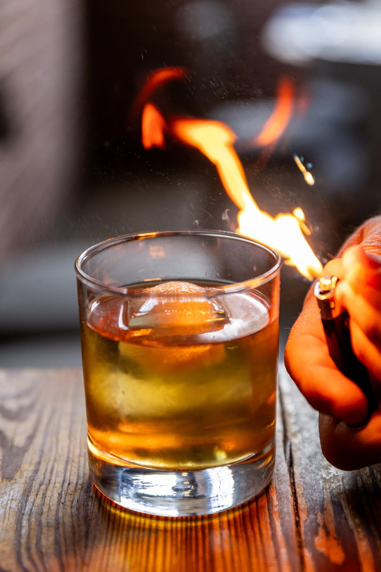 A person holding a lighter with a flame close to a glass of whiskey on a wooden table. Hot and Hot Fish Club Restaurant in Birmingham, Alabama.