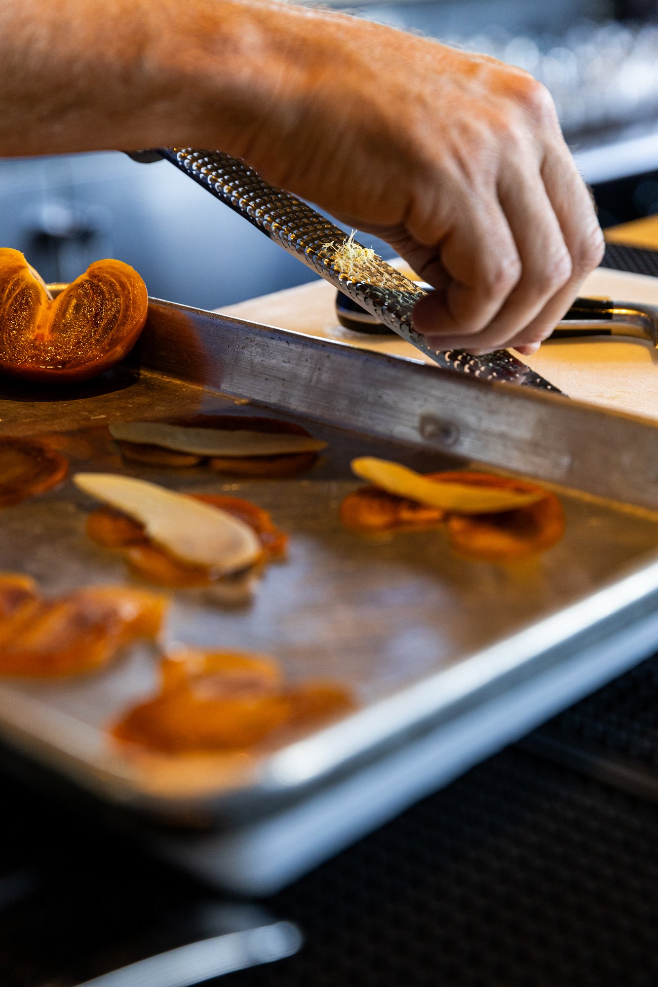 Person grating lemon zest over roasted tomato slices and garlic on a baking sheet. Hot and Hot Fish Club Restaurant in Birmingham, Alabama.