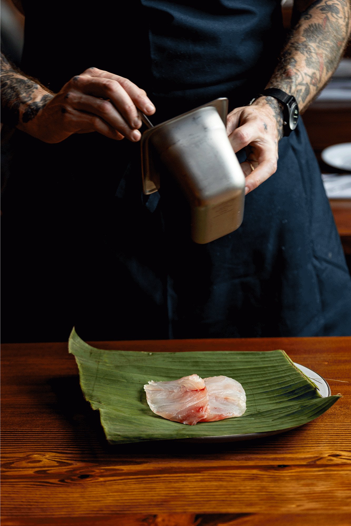 Person with tattooed arms and black watch seasoning a piece of raw fish on a banana leaf with a metal container.