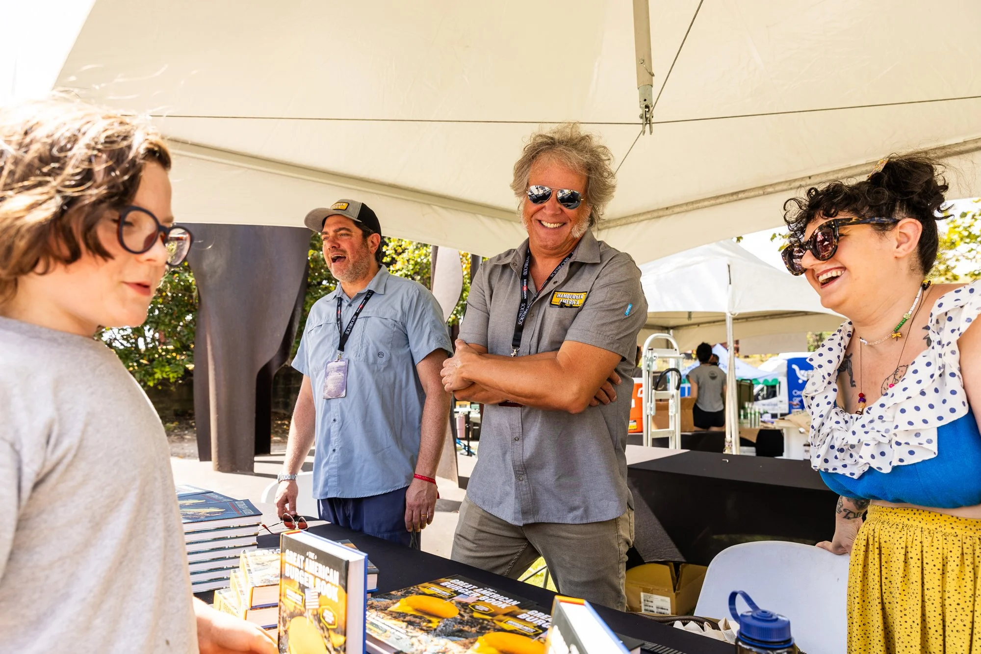 Four people at an outdoor booth under a white canopy, smiling and engaging with each other. Two women, one with glasses and a polka-dot top, and a man with gray hair and sunglasses, are talking with two men, one wearing a cap, and the other with curl