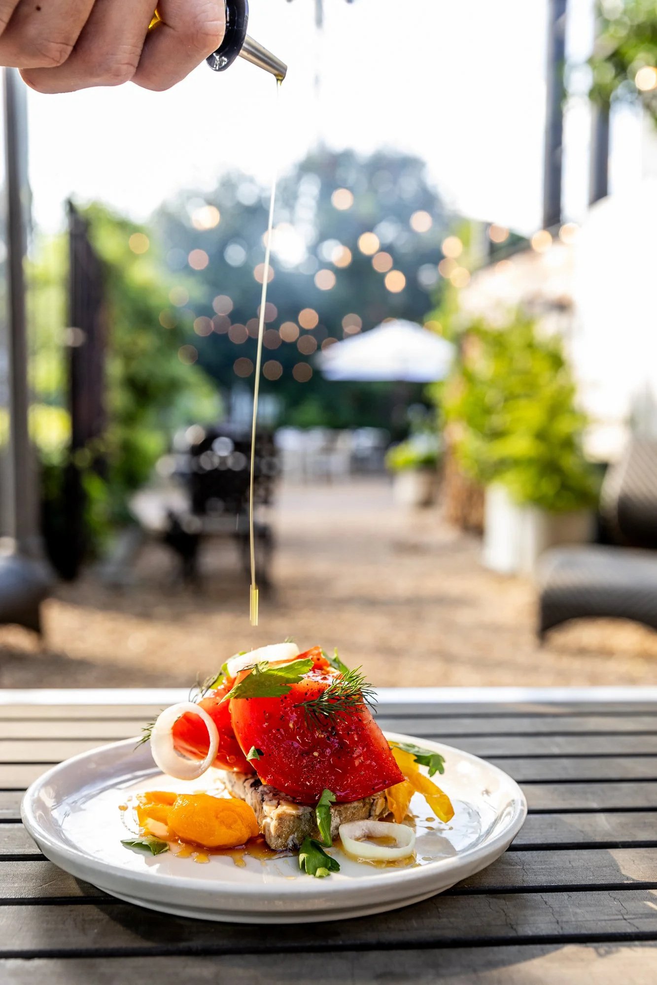 A plate of food on a wooden table with a hand pouring oil or dressing over the dish, outdoor setting with string lights, trees, and patio furniture in the background. Ovenbird Restaurant in Birmingham, Alabama.