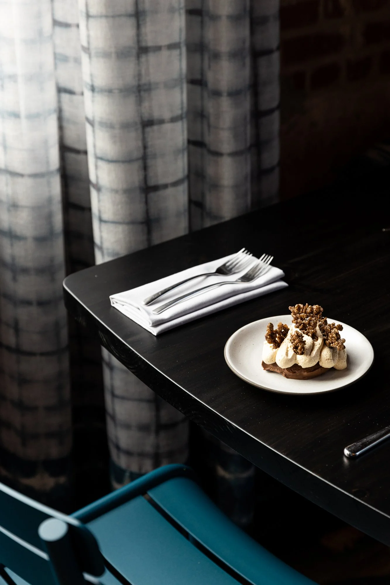A dessert on a white plate includes whipped cream and crumbly topping, placed on a dark wooden table next to neatly folded white napkins with forks, with curtains in the background.