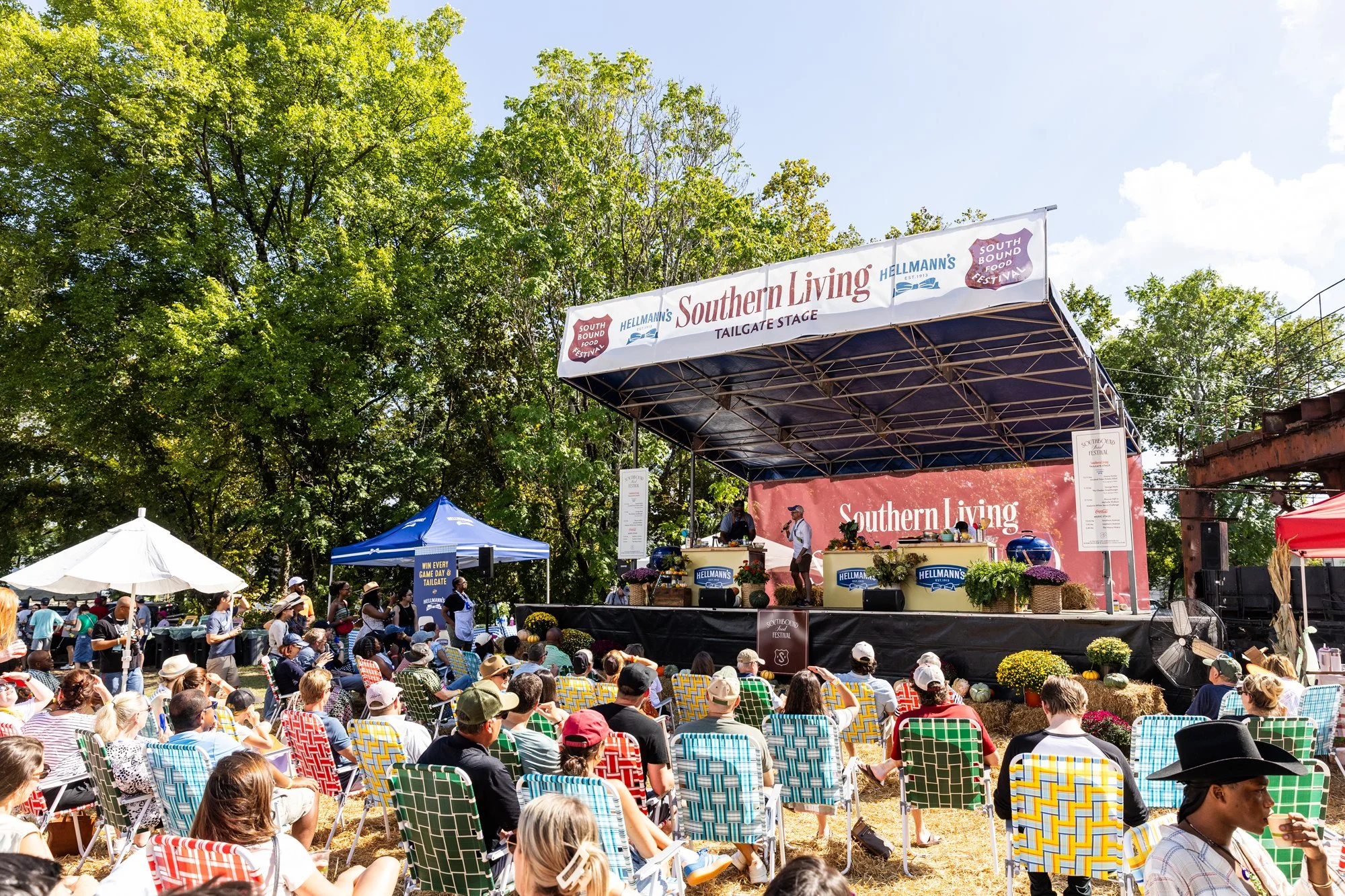 A large outdoor stage with a sign that reads "Southern Living Tailgate Stage" during the Southbound Food Festival, with a crowd of people seated on lawn chairs and standing, some wearing hats, under a partly cloudy sky surrounded by green trees.
