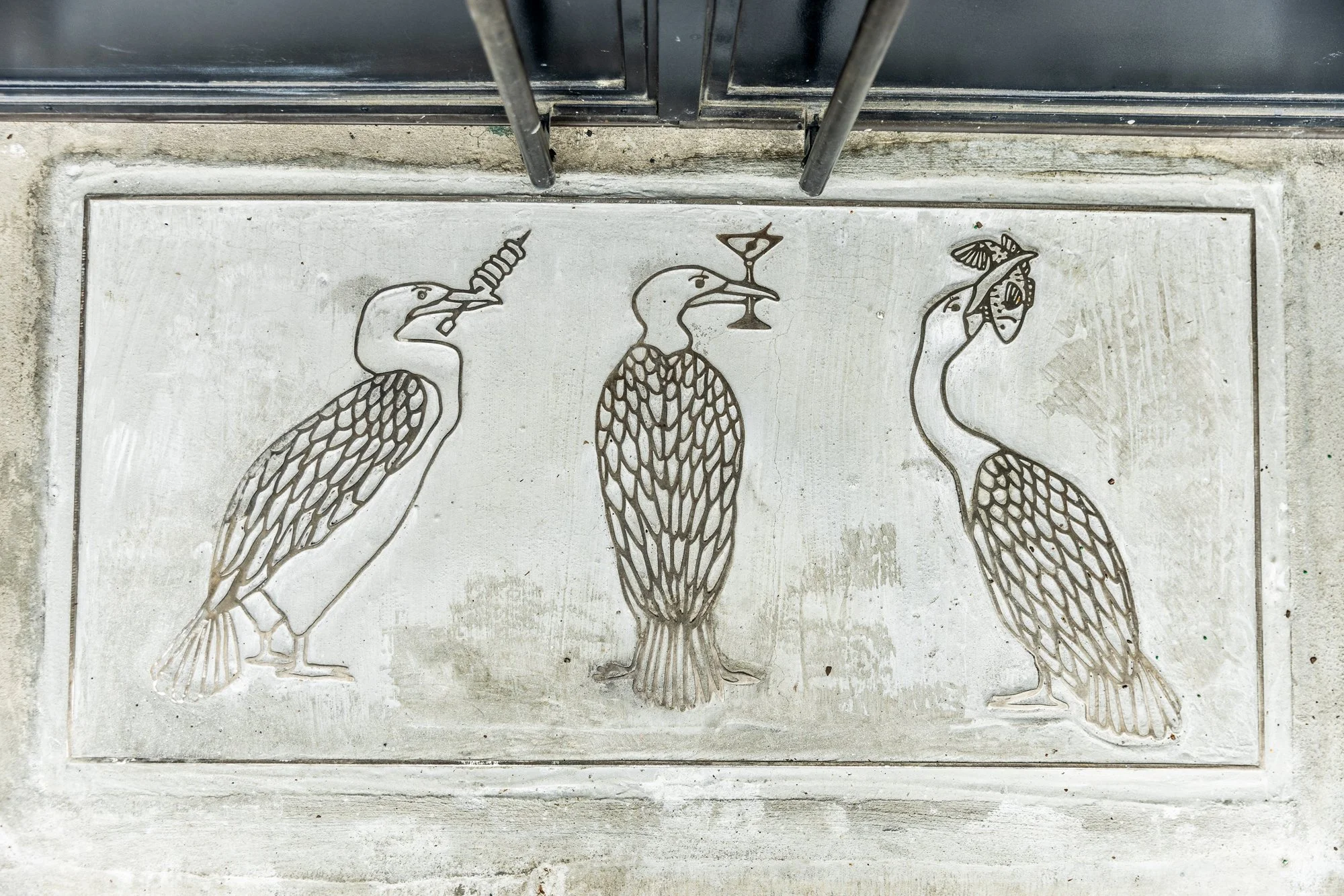 Decorative concrete sidewalk with three stylized bird illustrations, each holding different objects in their beaks: a skewer with a sausage, a martini glass, and a fish.