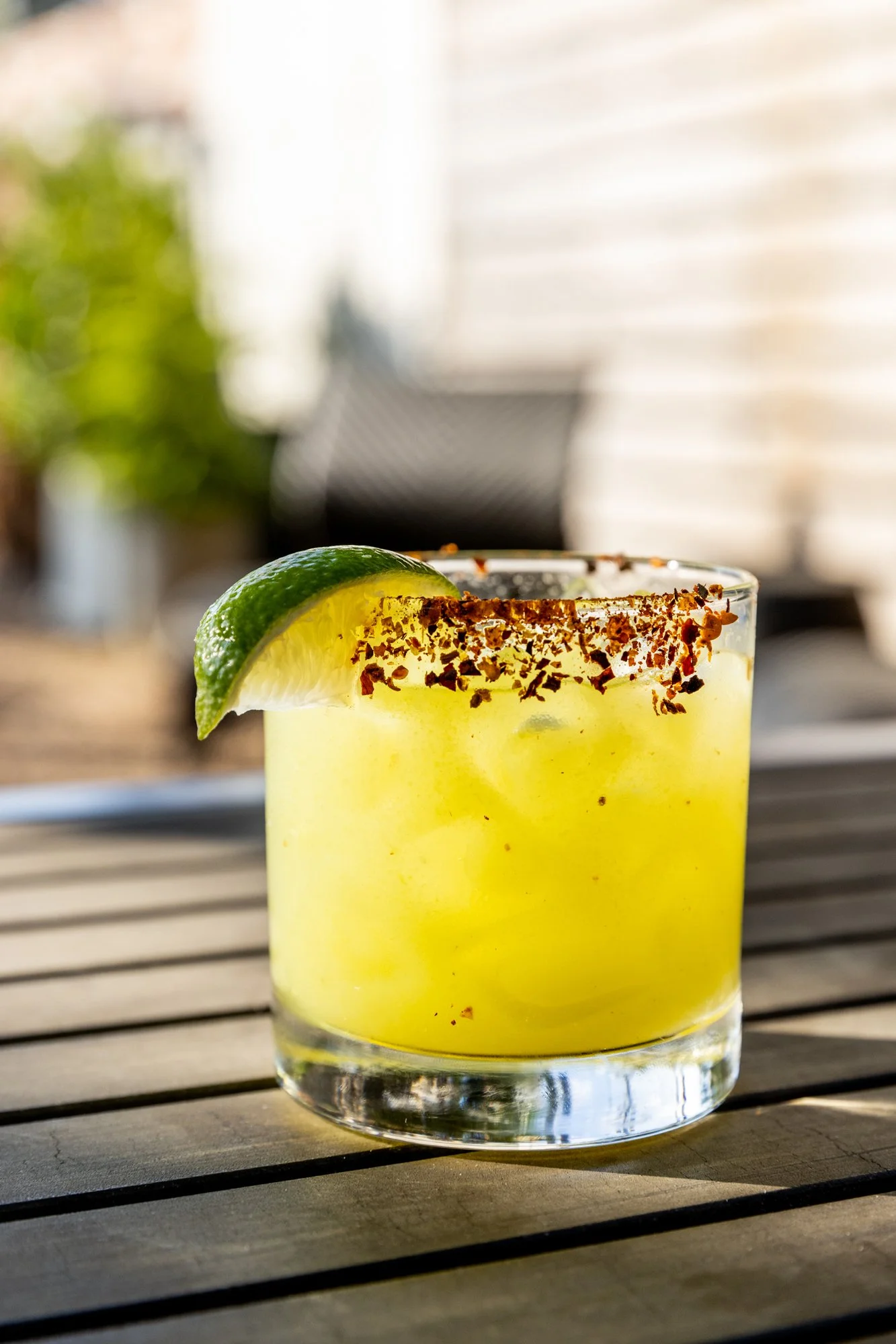 A yellow cocktail in a clear glass with a lime wedge garnish and chili powder rim, placed on a wooden surface outdoors. Ovenbird Restaurant in Birmingham, Alabama.