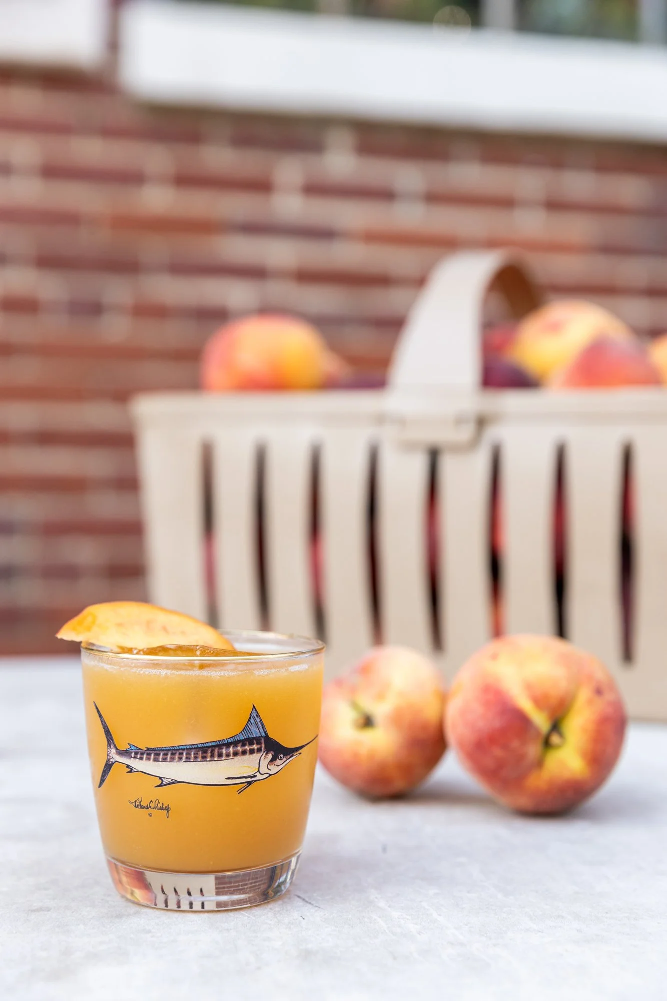 A glass of orange juice with a slice of peach on the rim, placed on a white surface with three peaches beside it. In the background, a white basket filled with more peaches is visible against a red brick wall. Hot and Hot Fish Club Restaurant