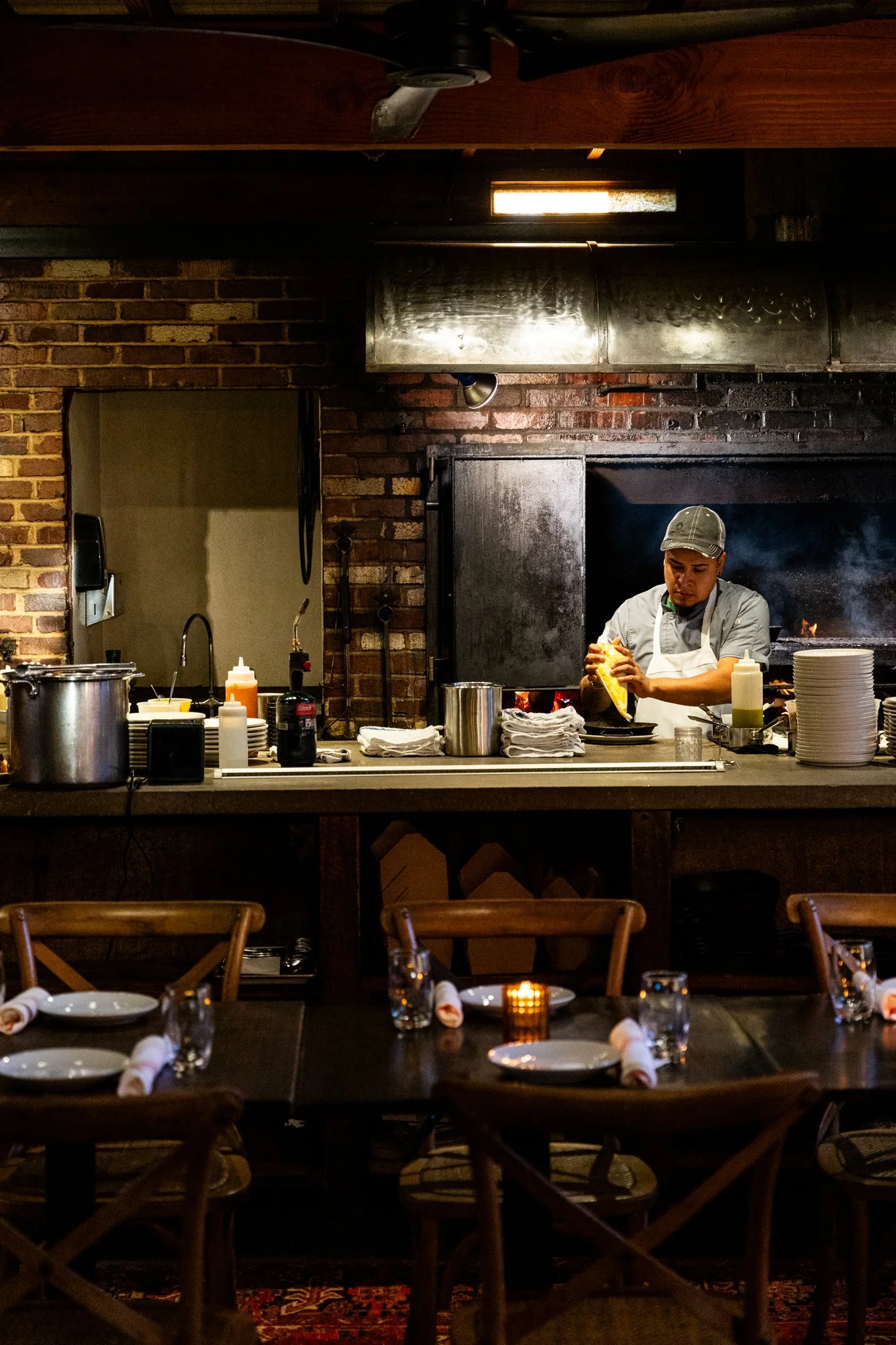 A chef prepares food in an open kitchen with brick walls, with tables set for dining in the foreground. Ovenbird Restaurant in Birmingham, Alabama.