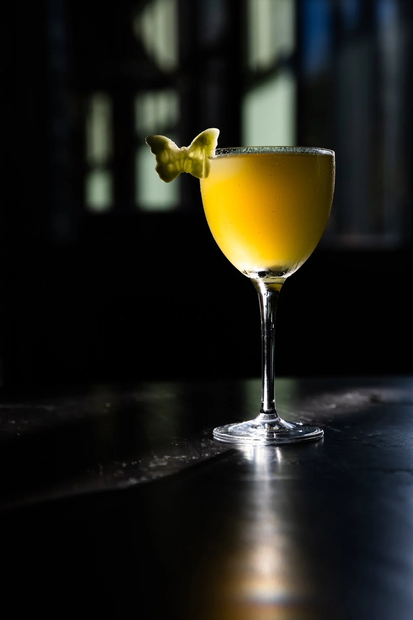 A yellow cocktail garnished with a butterfly-shaped piece of fruit, served in a tall, elegant glass with a long stem, on a dark surface with a blurred window in the background.