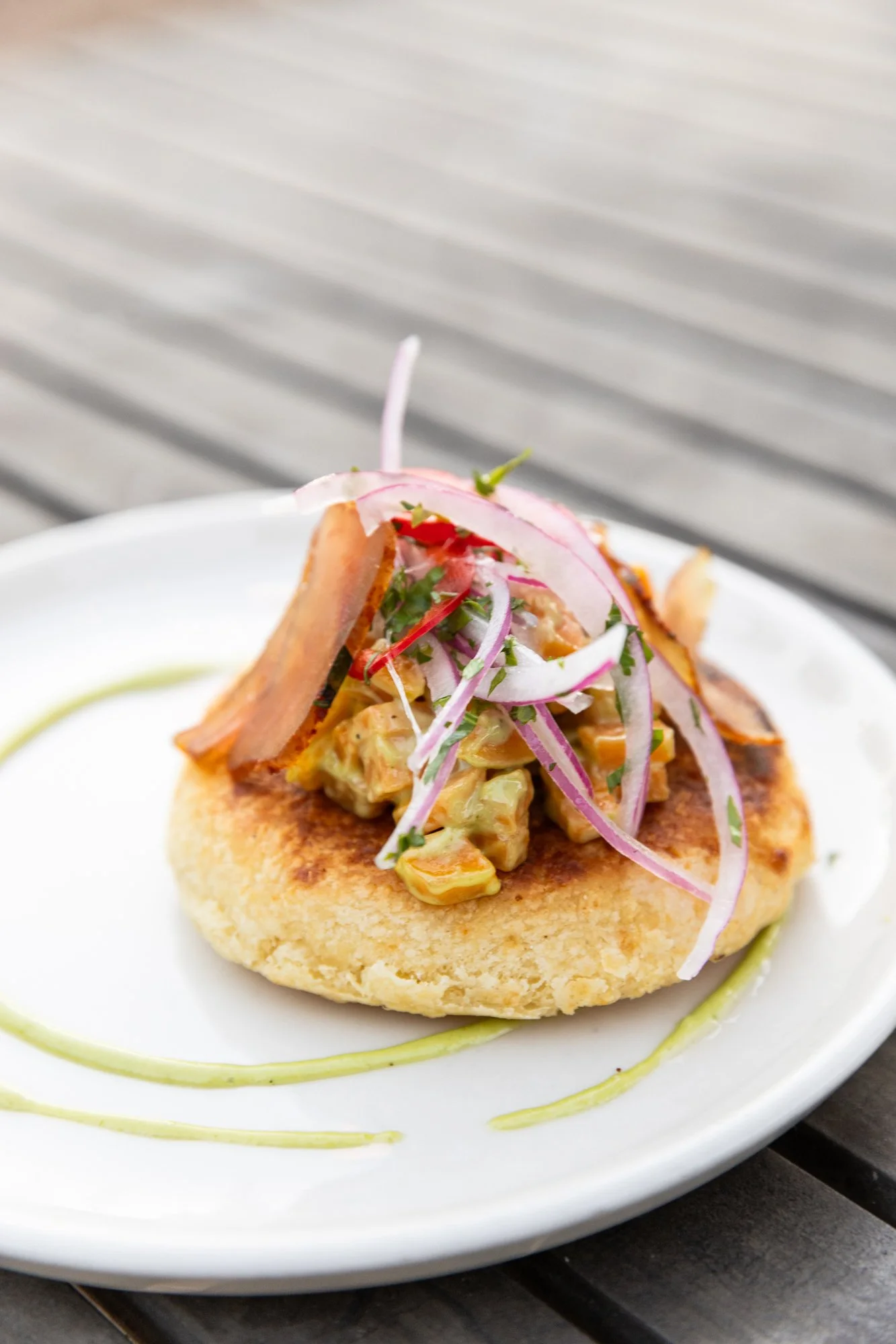 A serving of fish cake topped with a potato salad, garnished with sliced red onions, cilantro, and crispy bacon, on a white plate with green sauce drizzled around. Ovenbird Restaurant in Birmingham, Alabama.