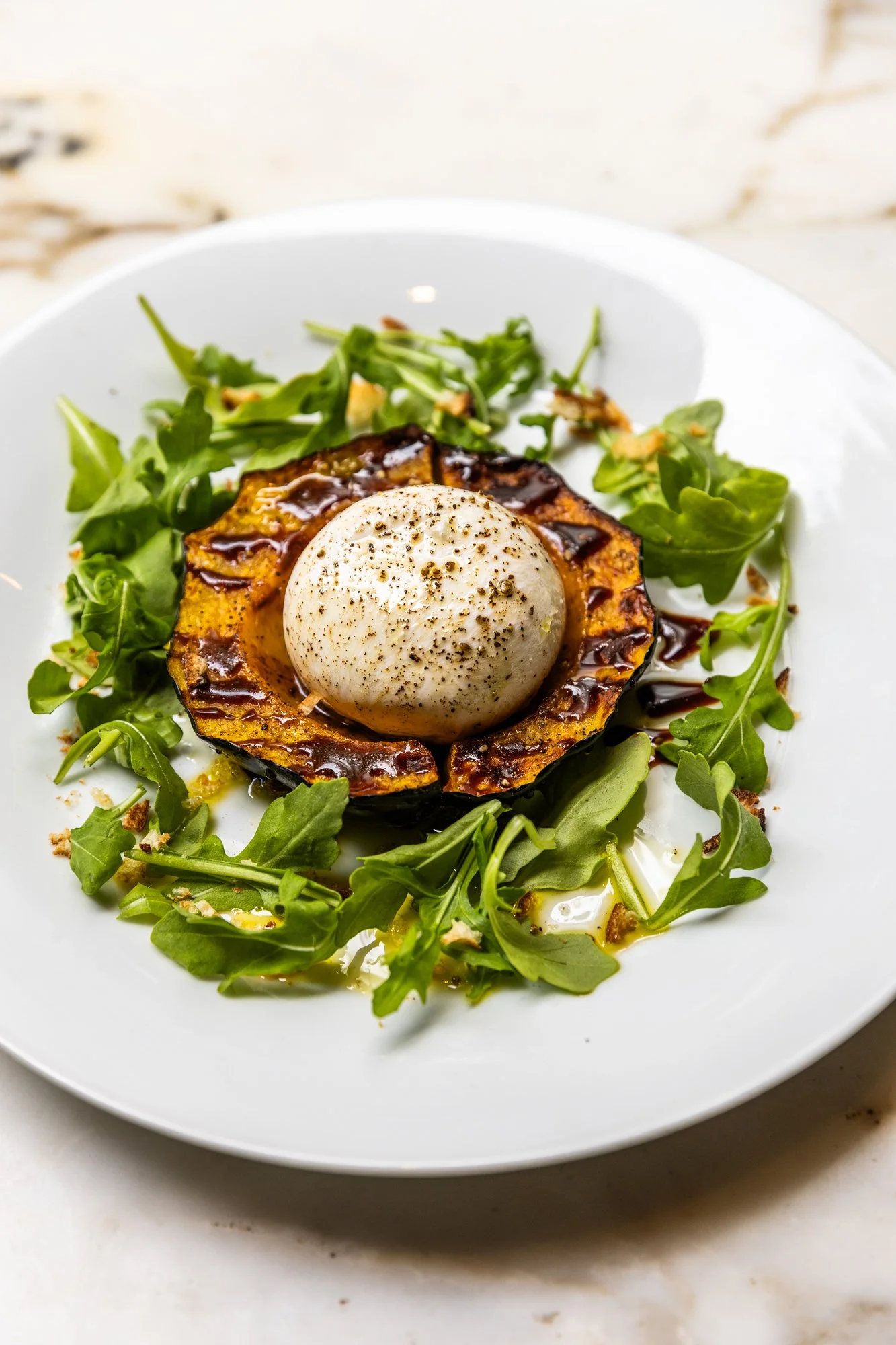 A plate with roasted eggplant topped with burrata cheese, surrounded by arugula leaves and drizzled with balsamic glaze. Ovenbird Restaurant in Birmingham, Alabama.