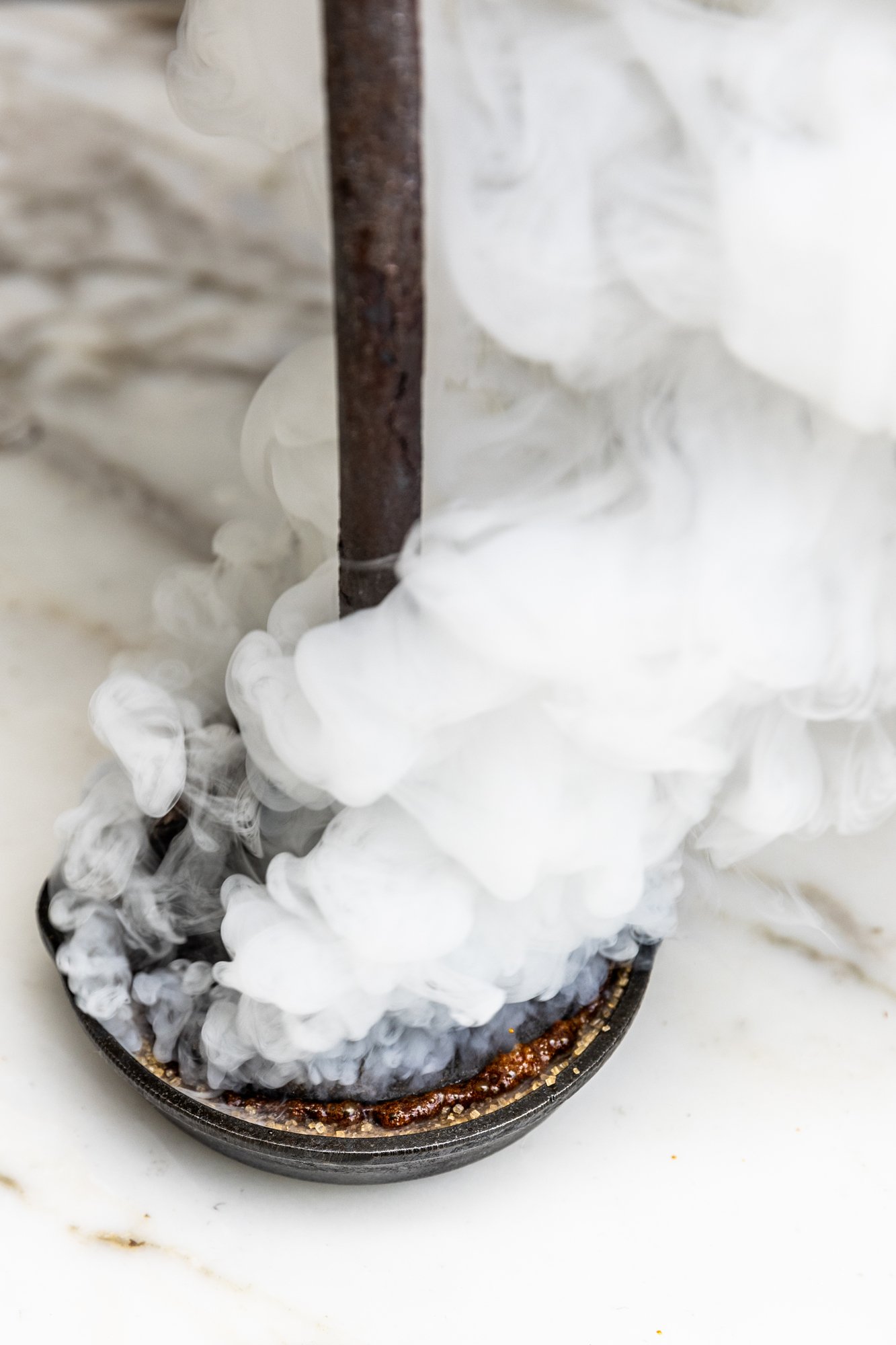A black skillet with what appears to be a spicy sauce at the bottom, with white vapor or smoke rising from it, and a rusted metal rod inserted in the center. Ovenbird Restaurant in Birmingham, Alabama.