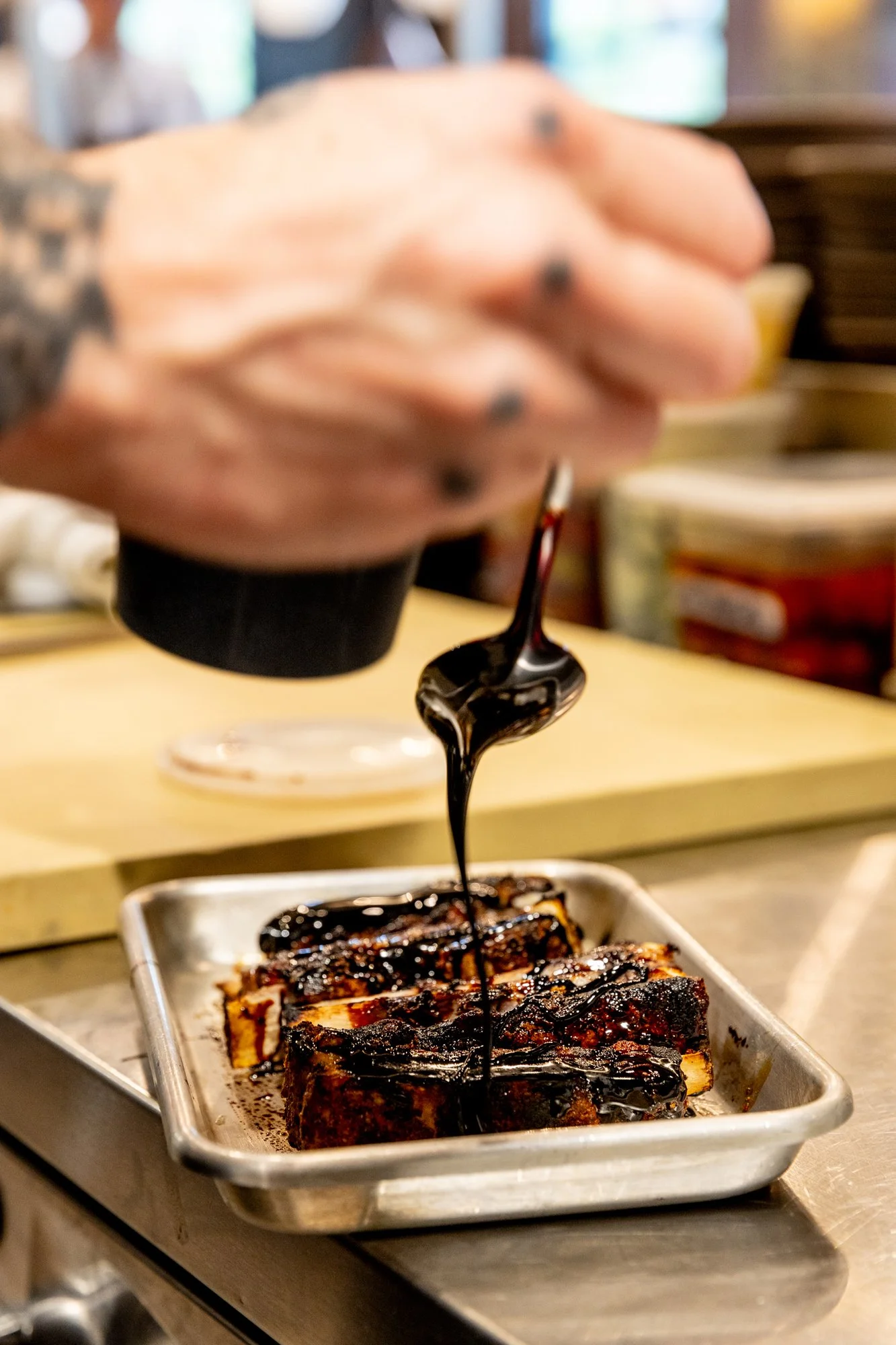A person drizzling dark barbecue sauce over grilled ribs on a metal tray in a kitchen or restaurant setting.