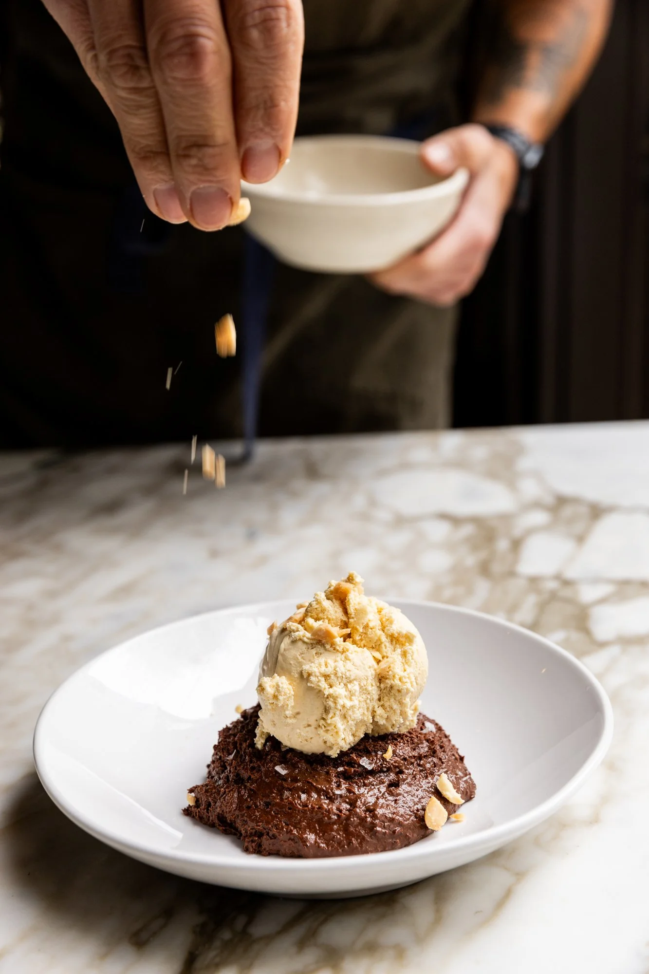 Person sprinkling a topping onto a dessert with chocolate cake and vanilla ice cream on a white plate. Ovenbird Restaurant in Birmingham, Alabama.