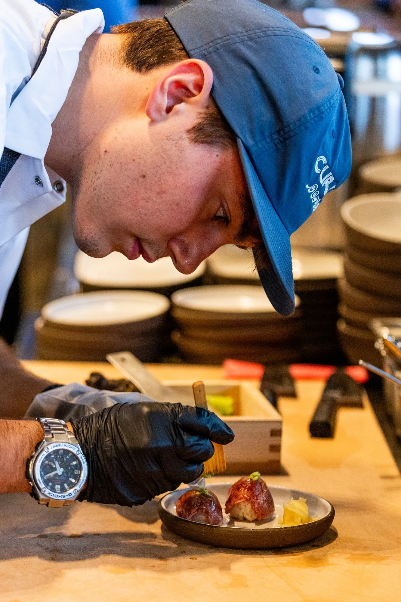 A young man wearing a blue cap and black gloves is carefully handling two sushi pieces with a brush, with yellow pickled ginger on a dish, in a restaurant kitchen setting.