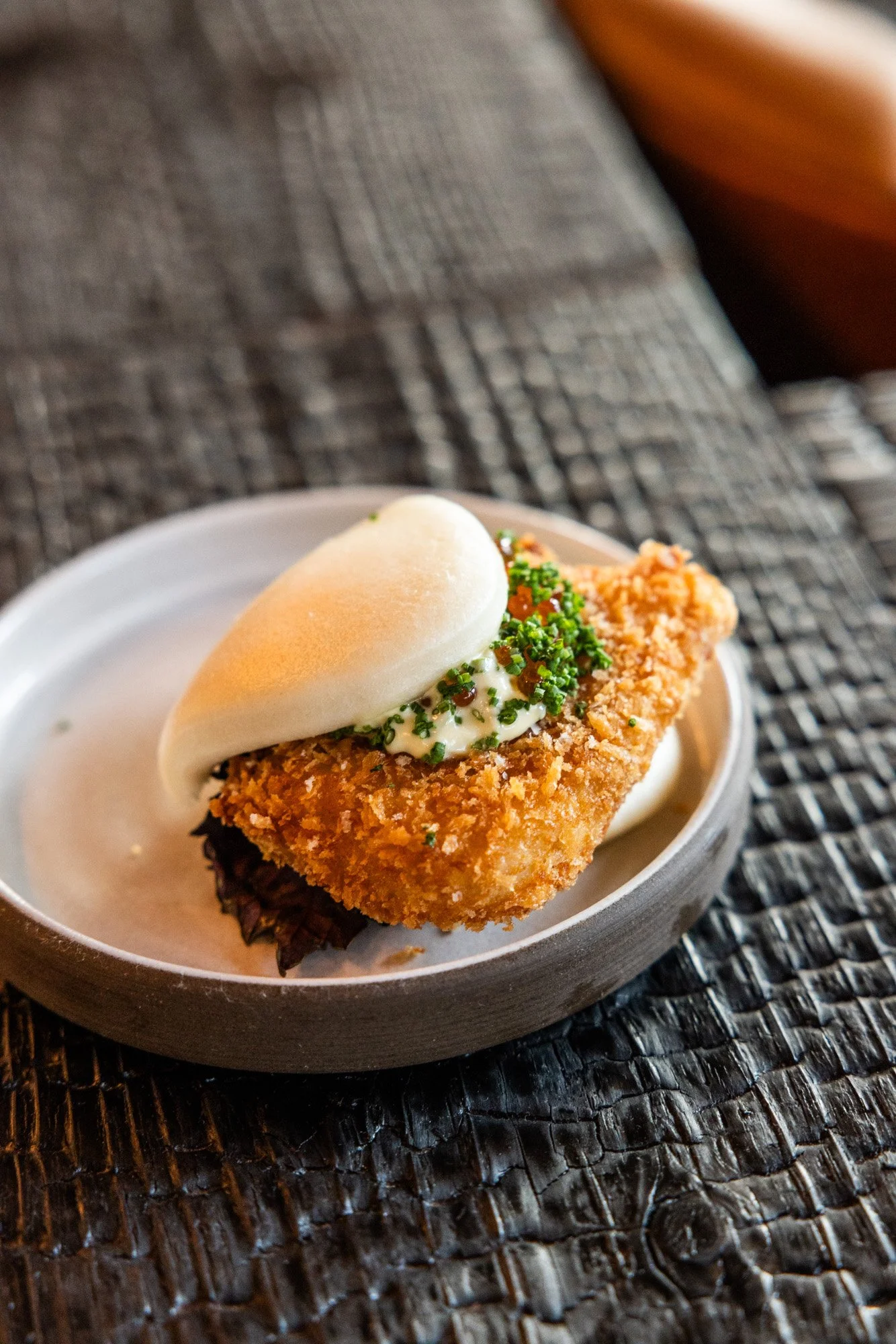 A breaded fried fish fillet topped with a dollop of tartar sauce and garnished with chopped chives, served on a small dish with a soft bread bun on top.