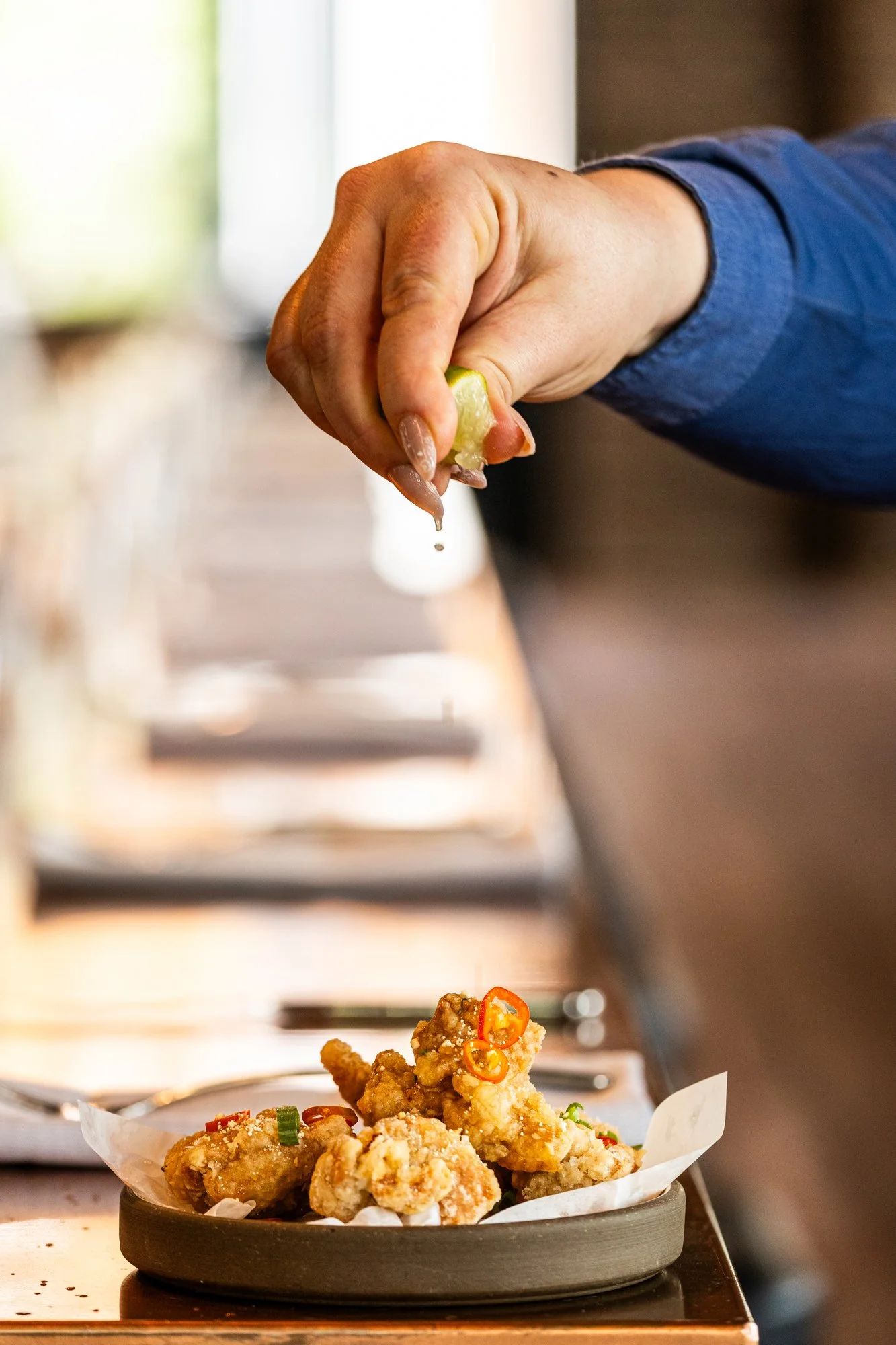 A person drizzling sauce on a plate of fried chicken bites topped with sliced red chili and green onions.