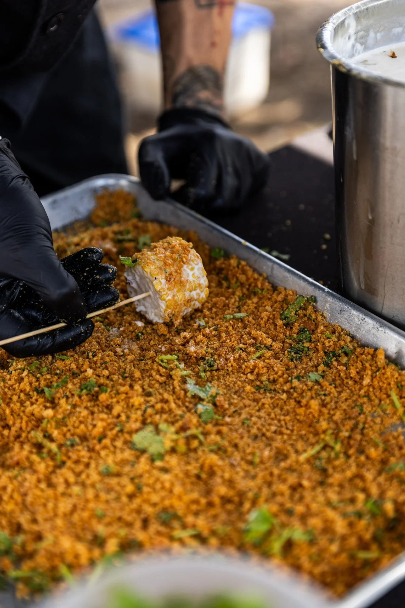 Person wearing black gloves placing a piece of cooked food on a skewer over a tray of seasoned rice or grains. The scene appears to be food preparation in an outdoor or commercial setting. Southbound Food Festival in Birmingham, Alabama.