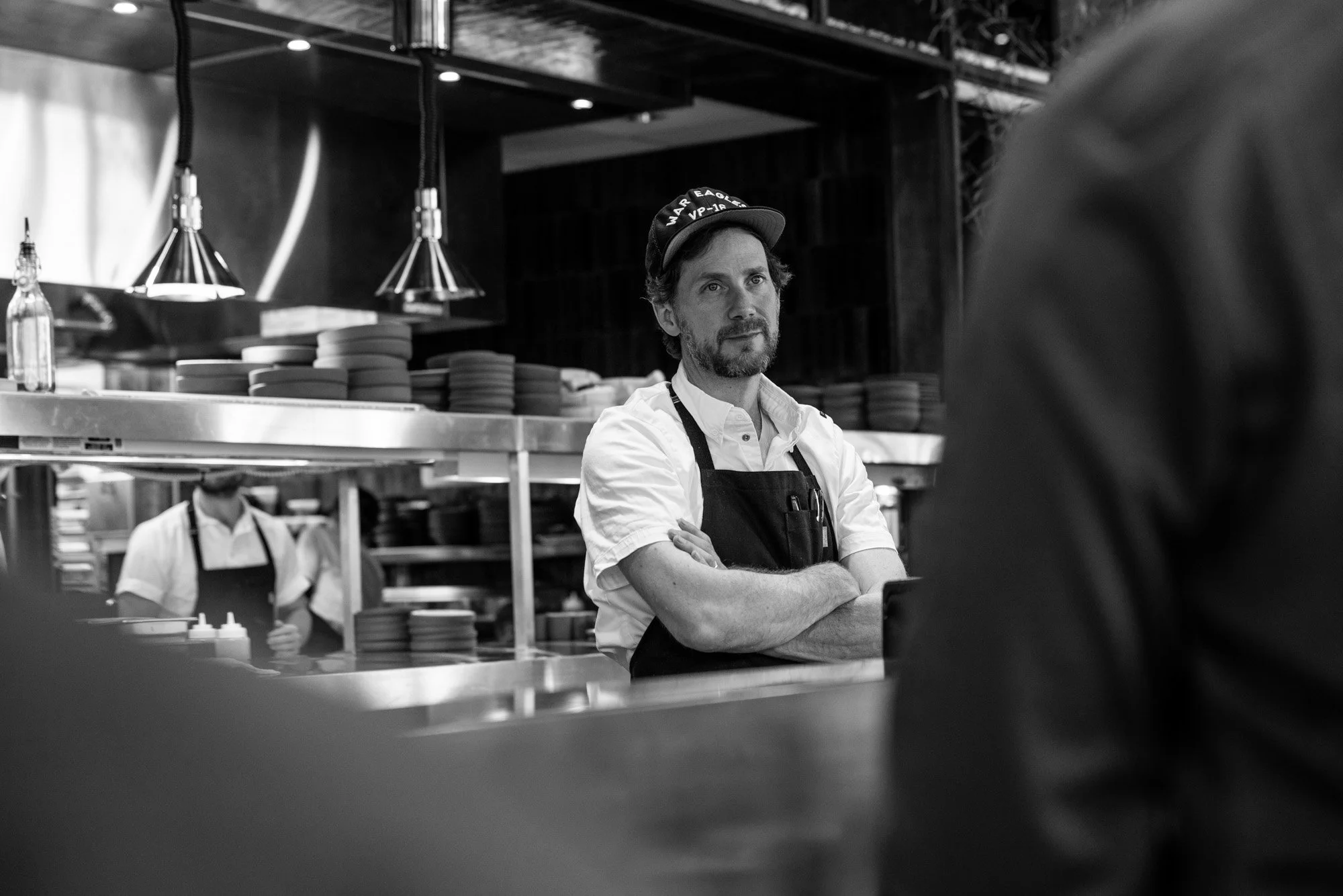 A man with a beard and mustache wearing a hat, white shirt, and apron stands with his arms crossed in a restaurant kitchen. There are plates on shelves and other kitchen staff in the background.