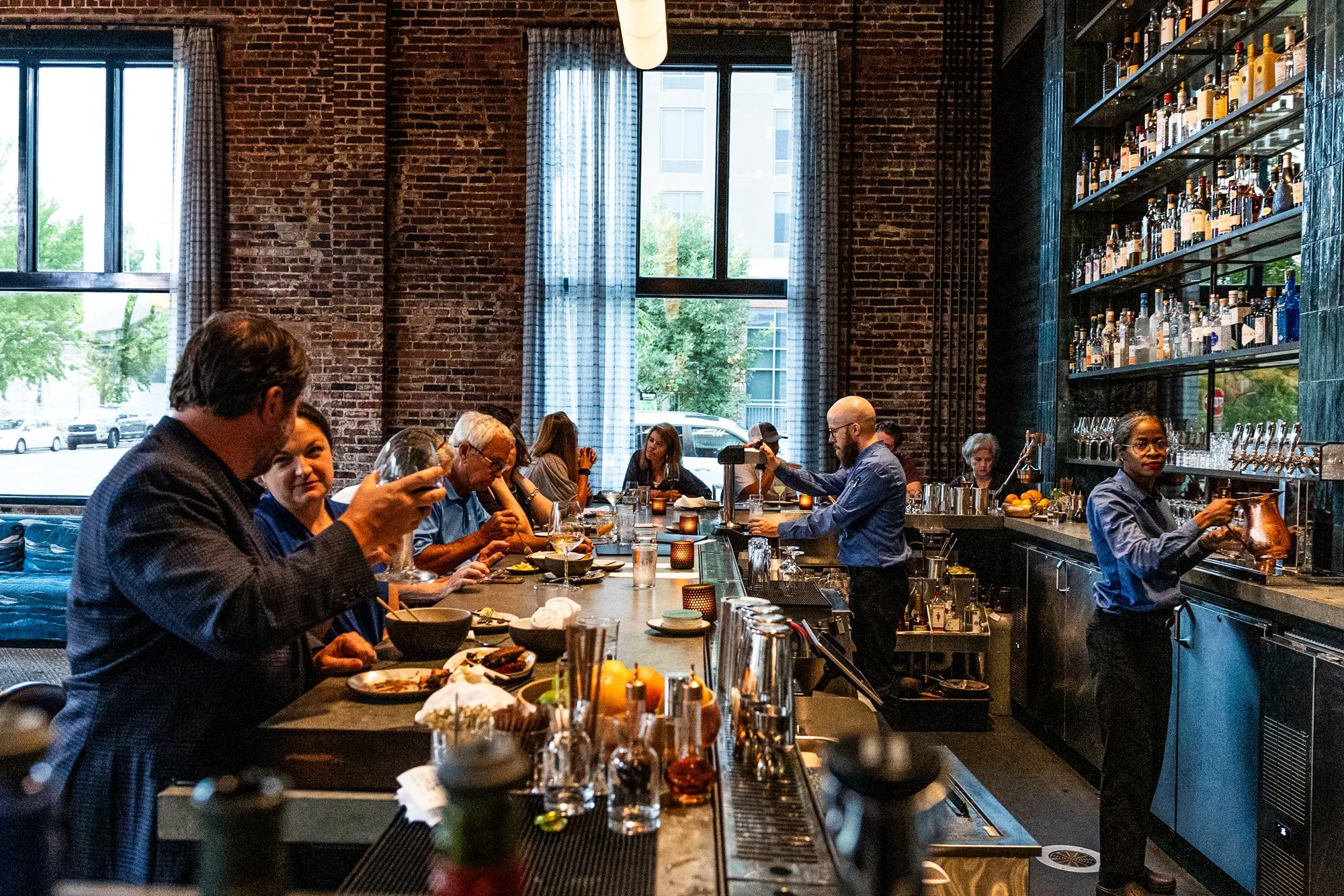 A busy restaurant or bar with patrons seated at a long counter, some eating and drinking, and bartenders preparing drinks behind the bar with bottles on shelves.