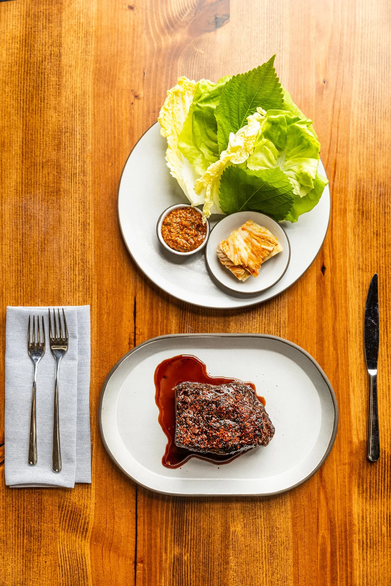 A plate with a cooked beef steak topped with sauce, and a side of leafy greens with dipping sauces on a wooden table.