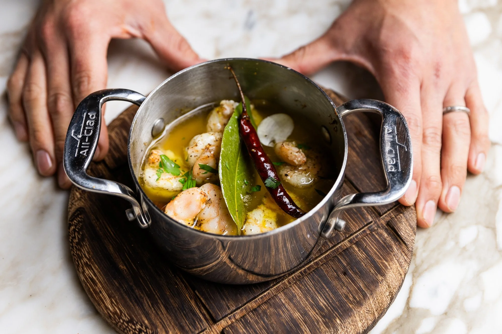 A small metal pot containing cooked shrimp, a bay leaf, a red chili pepper, and garlic, sitting on a round wooden cutting board with a person's hands framing it. Ovenbird Restaurant in Birmingham, Alabama.