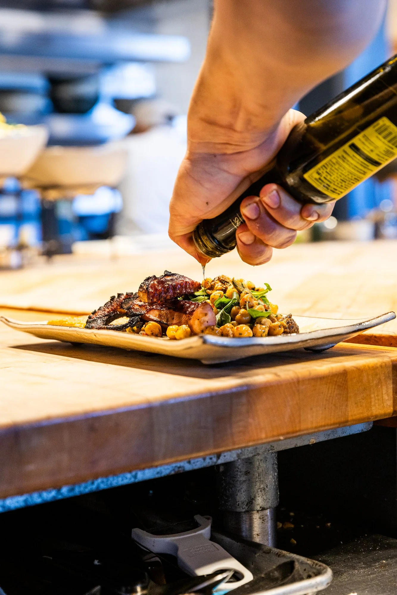 Person pouring sauce on a plated dish with bacon, chickpeas, and greens in a restaurant kitchen. Hot and Hot Fish Club Restaurant in Birmingham, Alabama.