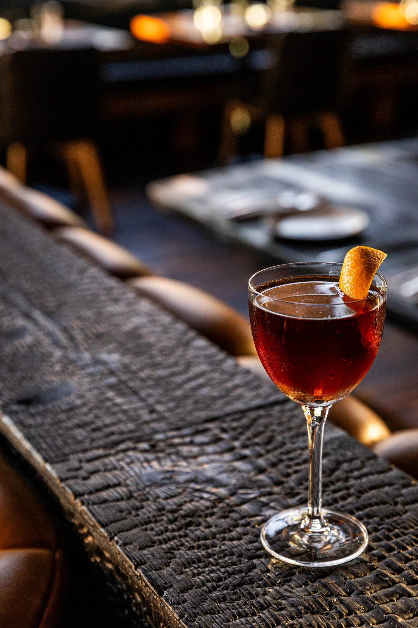 A cocktail in a wine glass with an orange peel garnish on a rustic wooden bar or table.