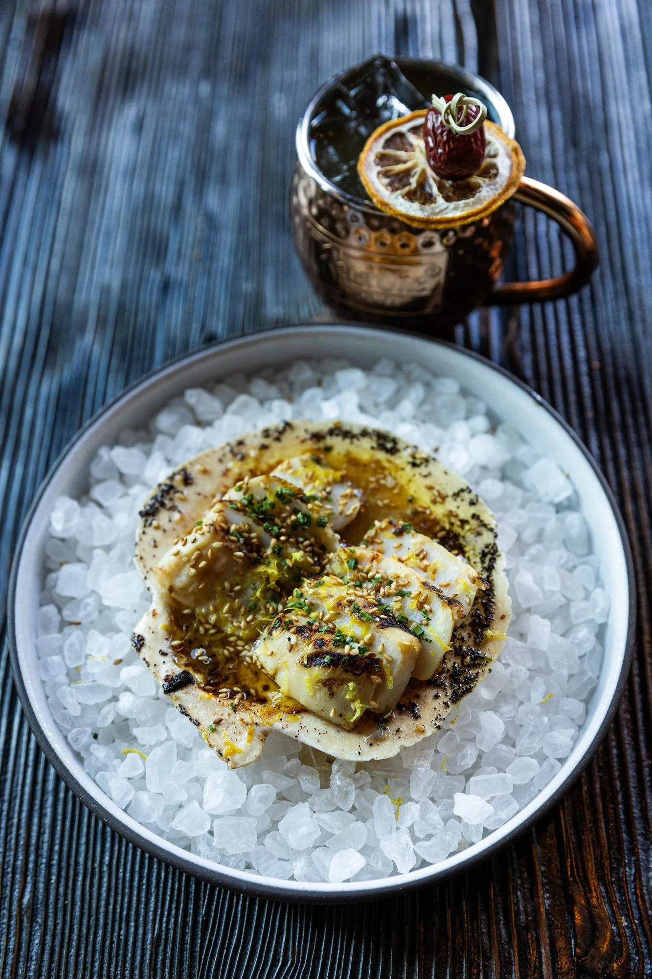 A dish of grilled octopus served on crushed ice with seasoning, garnished with lemon zest and chopped herbs, alongside a glass of dark soda with lemon and cherry garnish.