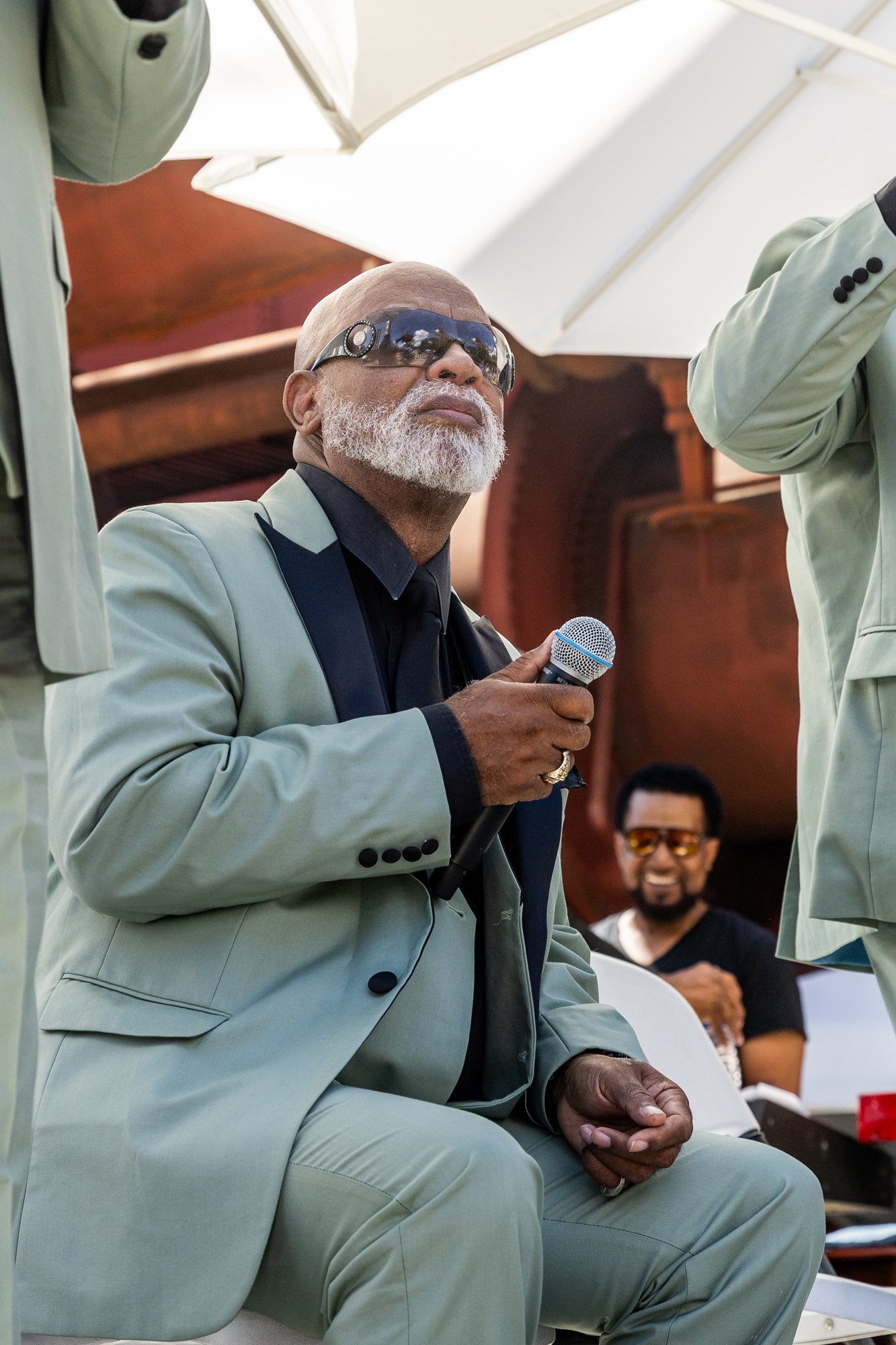 An older man with gray beard wearing sunglasses, a black shirt, and a gray suit, holding a microphone outdoors during a sunny day, with a person smiling in the background. Southbound Food Festival in Birmingham, Alabama.